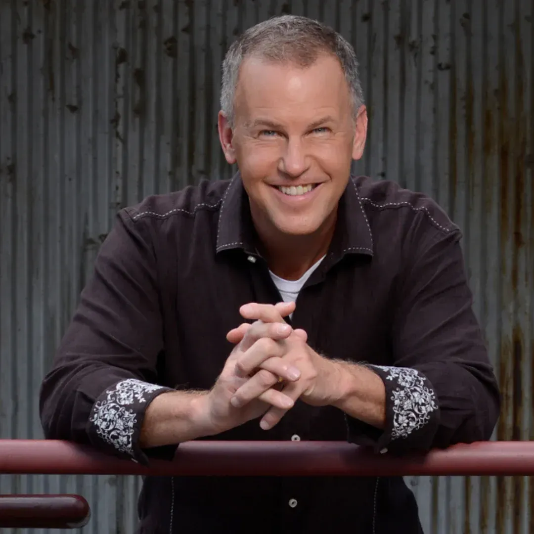 A smiling man leaning on a railing with his hands folded
