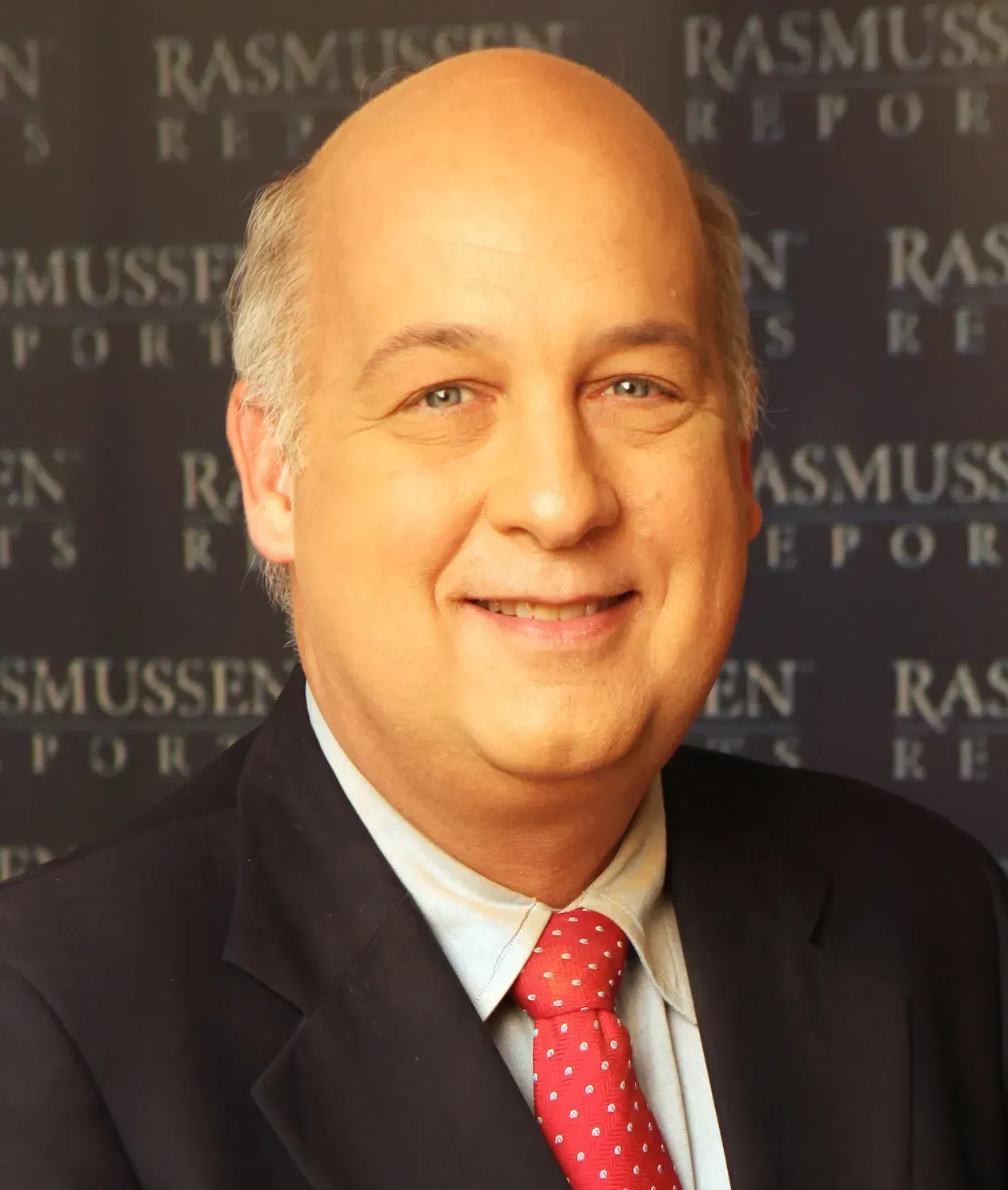 A man in a suit and tie is smiling in front of a sign that says rasmuss report