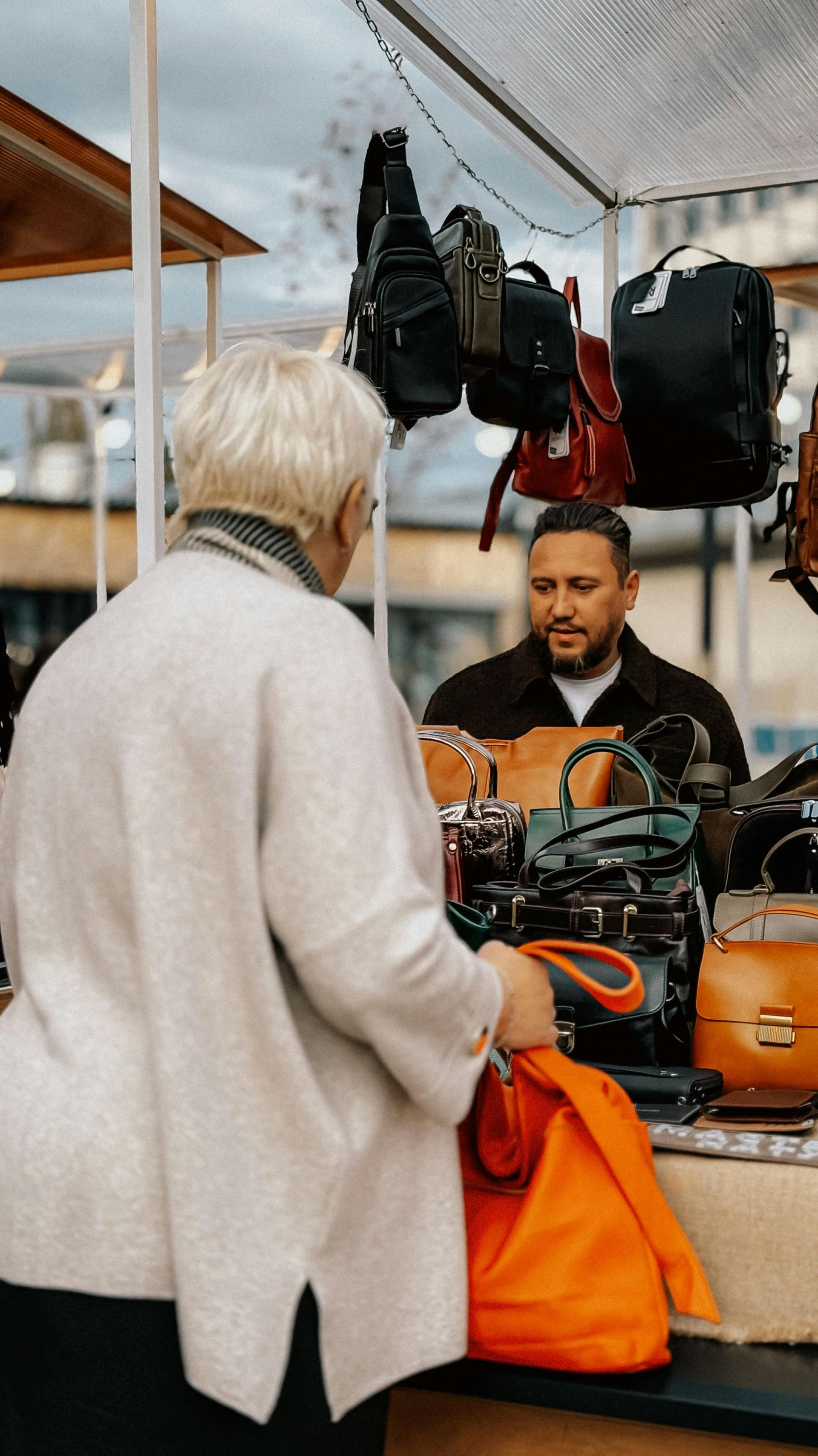 Person browsing accessories at an outdoor market stall with hanging bags and an orange tote.