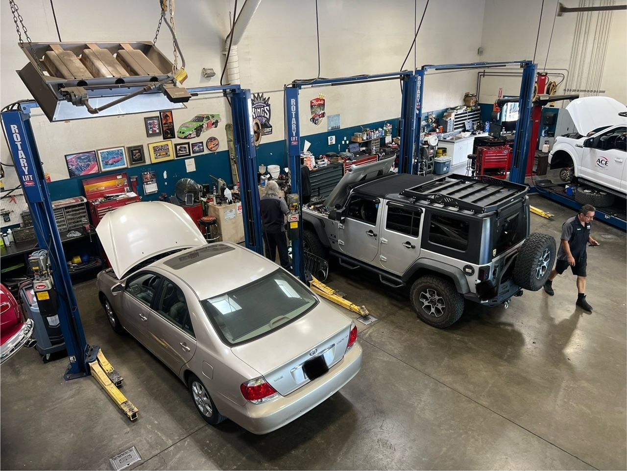 Auto repair shop with a silver sedan and gray Jeep on lifts, a mechanic working nearby.