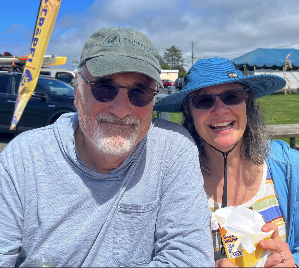 Smiling couple in hats and sunglasses at an outdoor event, with tents and flags in the background.