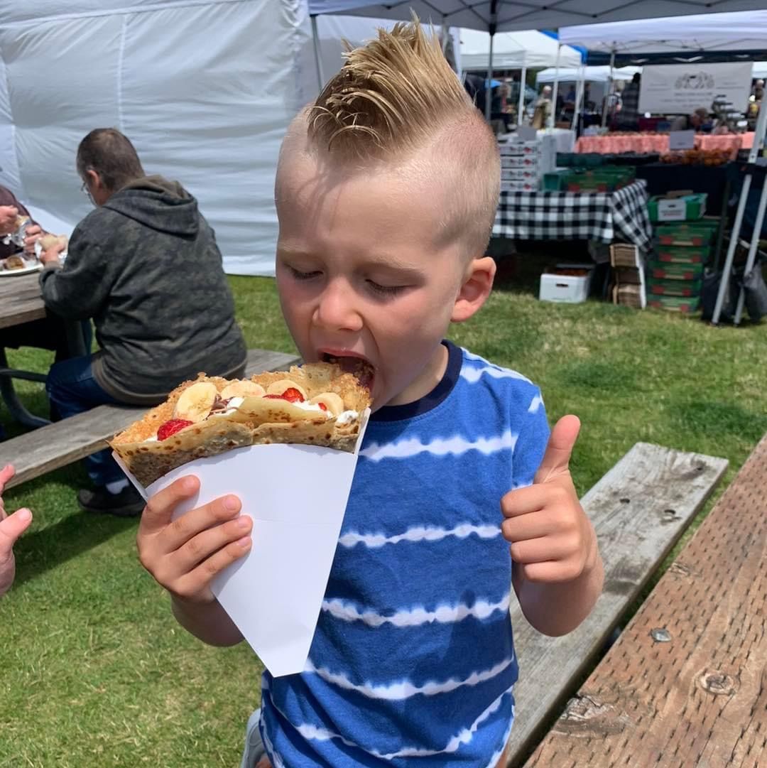 Child in a blue striped shirt eating a large crepe at an outdoor market booth