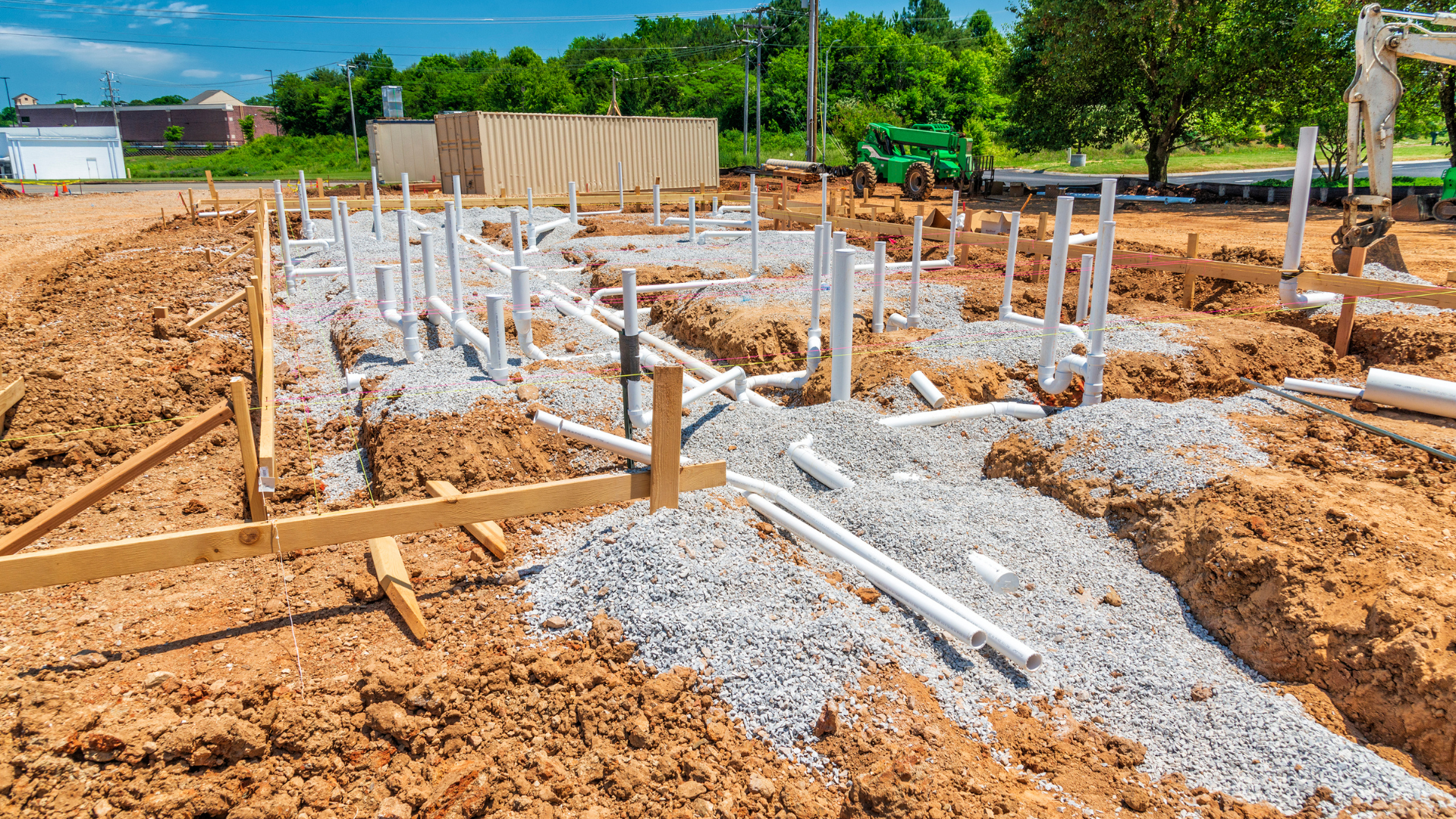 Construction site with foundation footings, rebar, white PVC pipes, and dirt under a clear blue sky