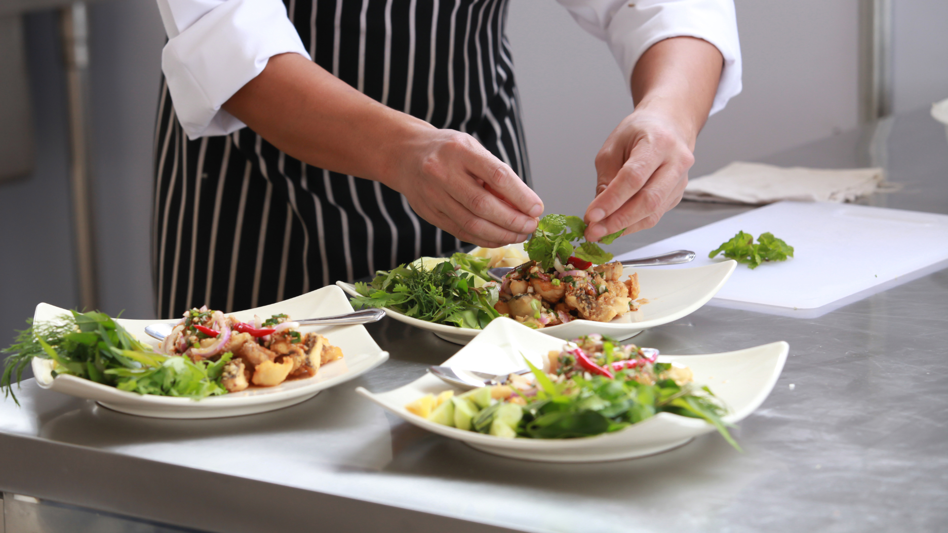 Chef plating salads on a stainless steel counter in a bright kitchen