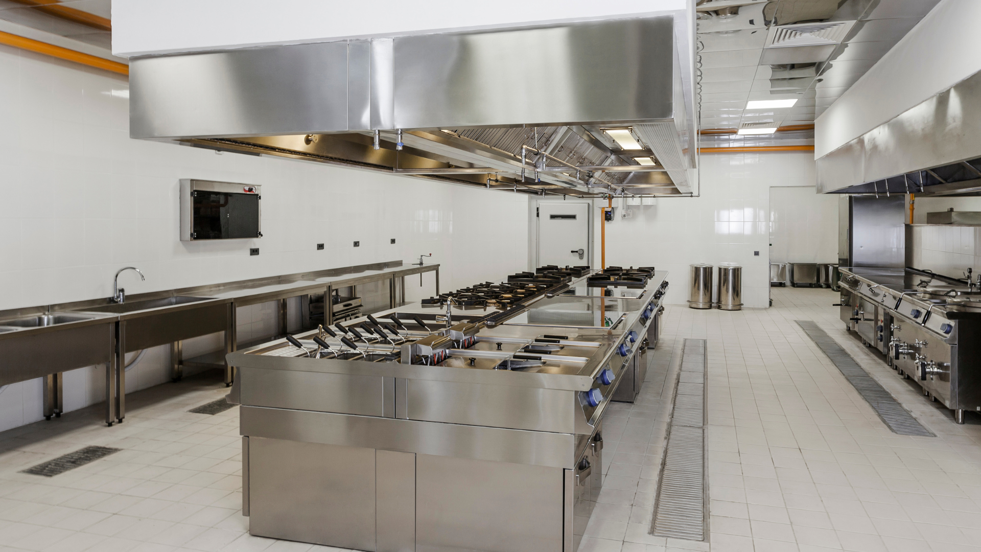 Empty stainless-steel commercial kitchen with rows of counters, sinks, and large overhead vents
