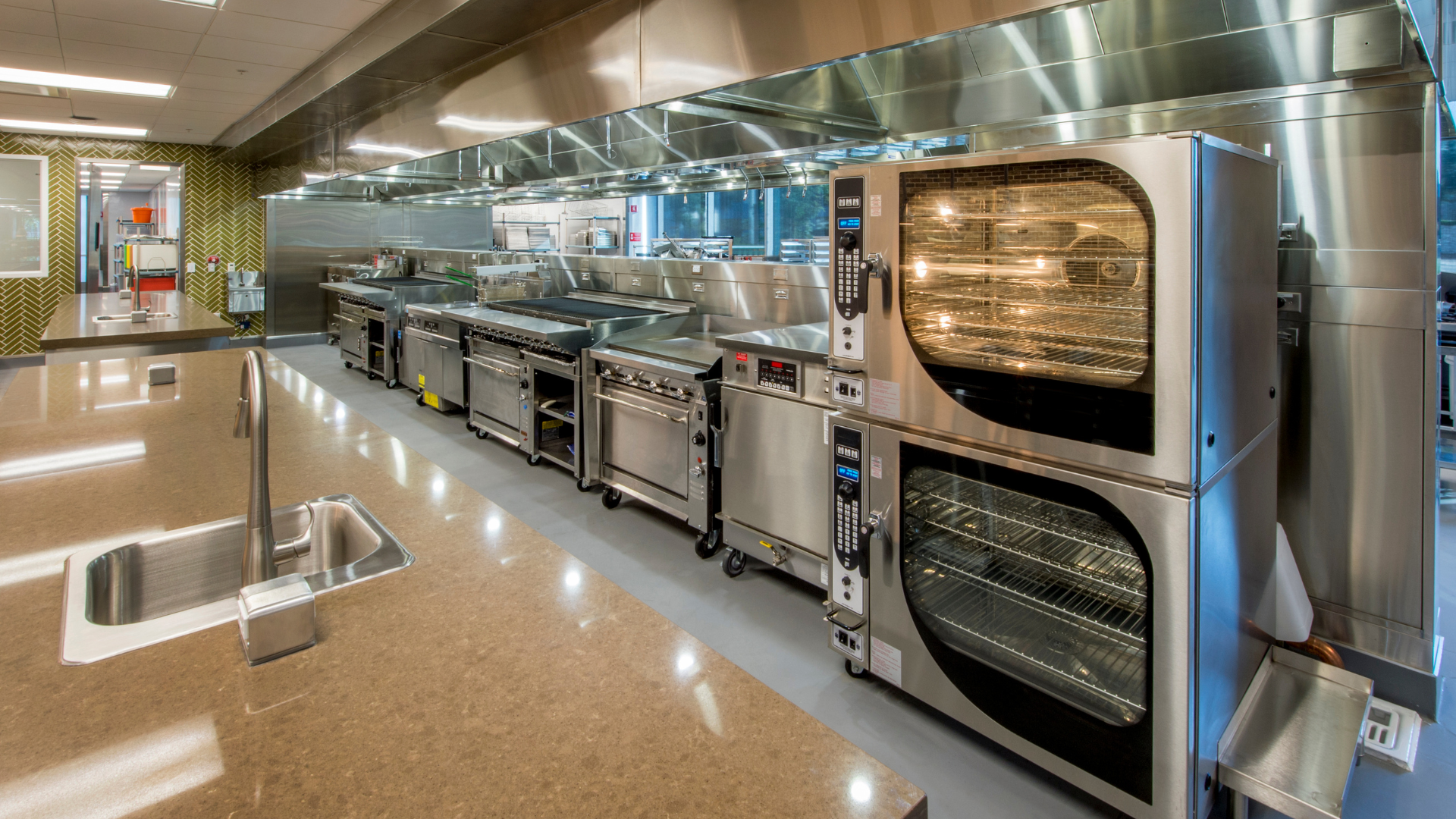 Commercial kitchen with stainless steel ovens and ranges under bright lights, clean polished floor