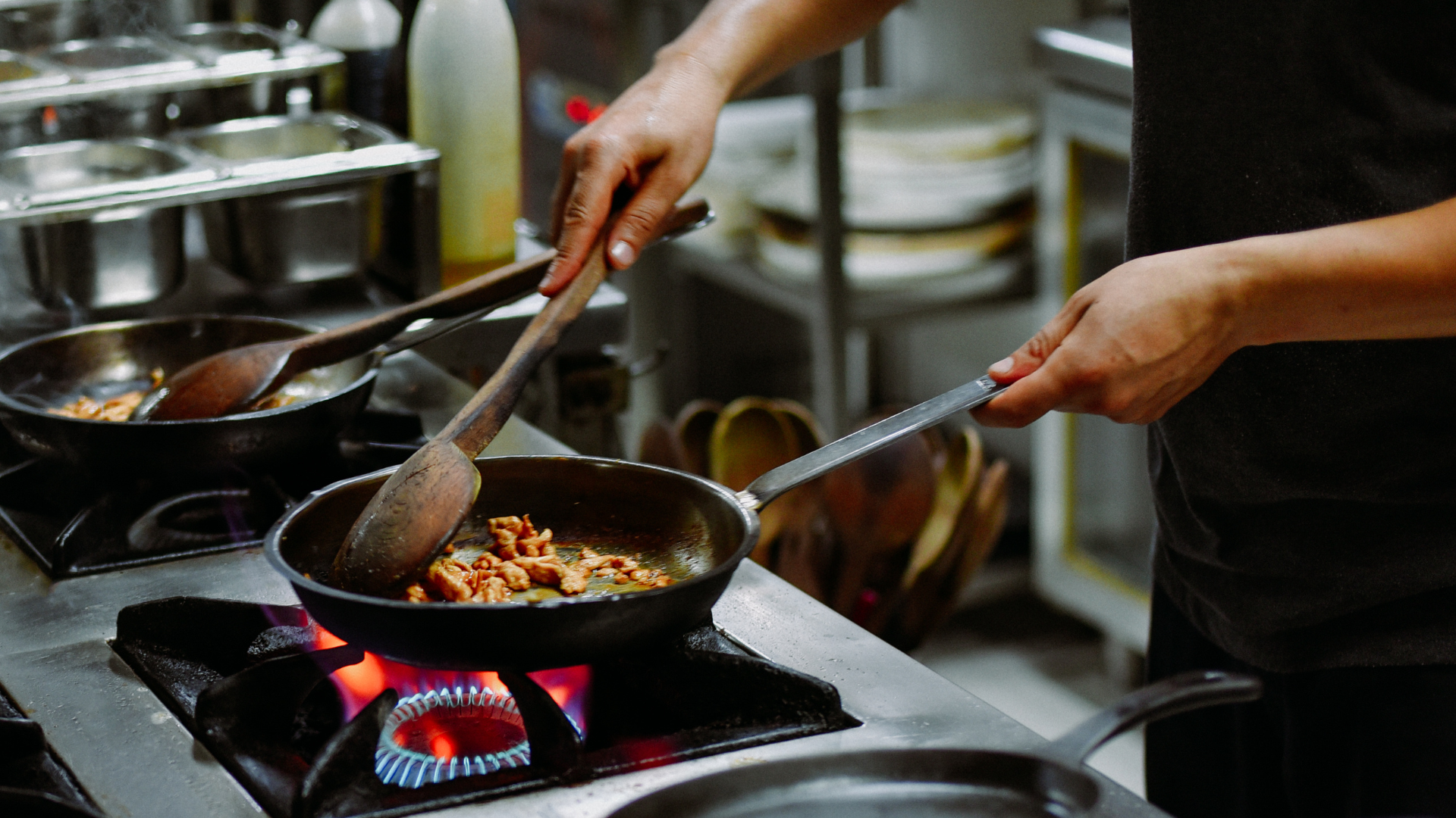 Two cooks stirring food in a pan over a gas stove in a busy kitchen