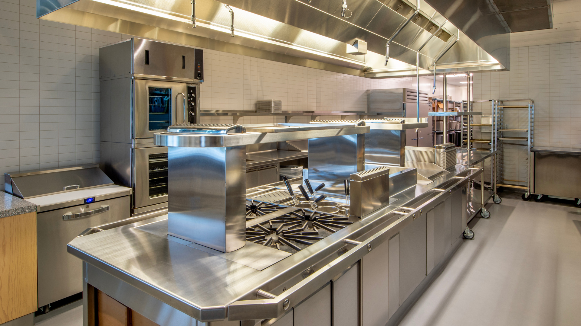 Stainless steel commercial kitchen with cooking stations, counters, and overhead ventilation in bright lighting