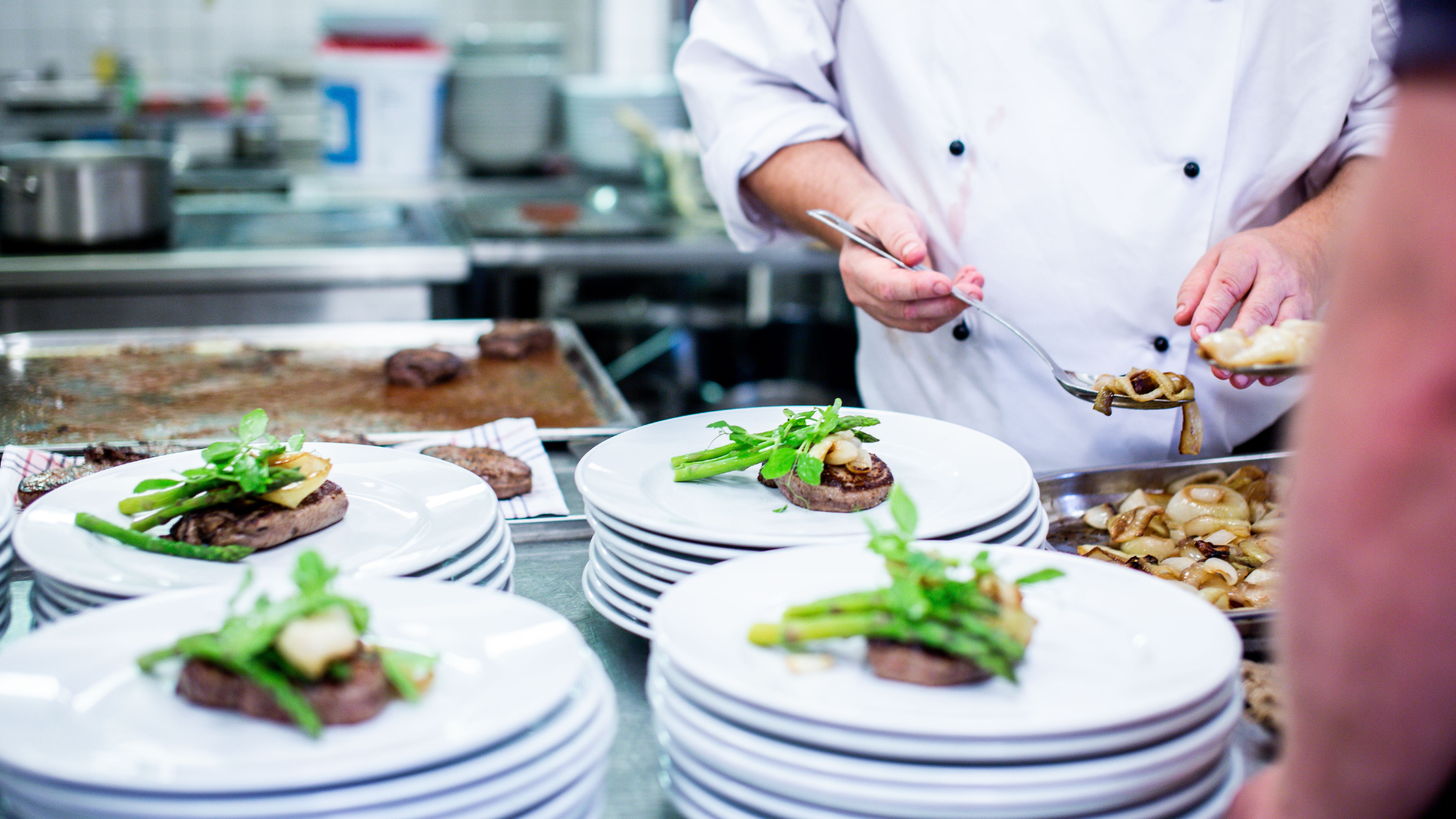 Chef plating green-topped dishes on stacked white plates in a kitchen