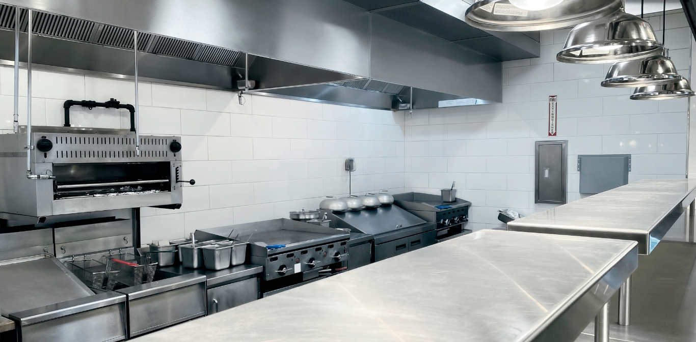 Empty stainless-steel commercial kitchen with counters, stoves, sinks, and hanging heat lamps