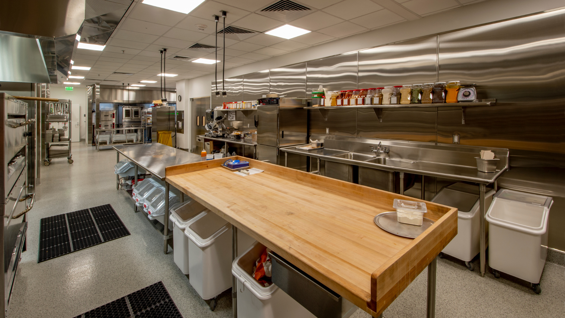 Commercial kitchen with stainless steel equipment and large wooden worktable.