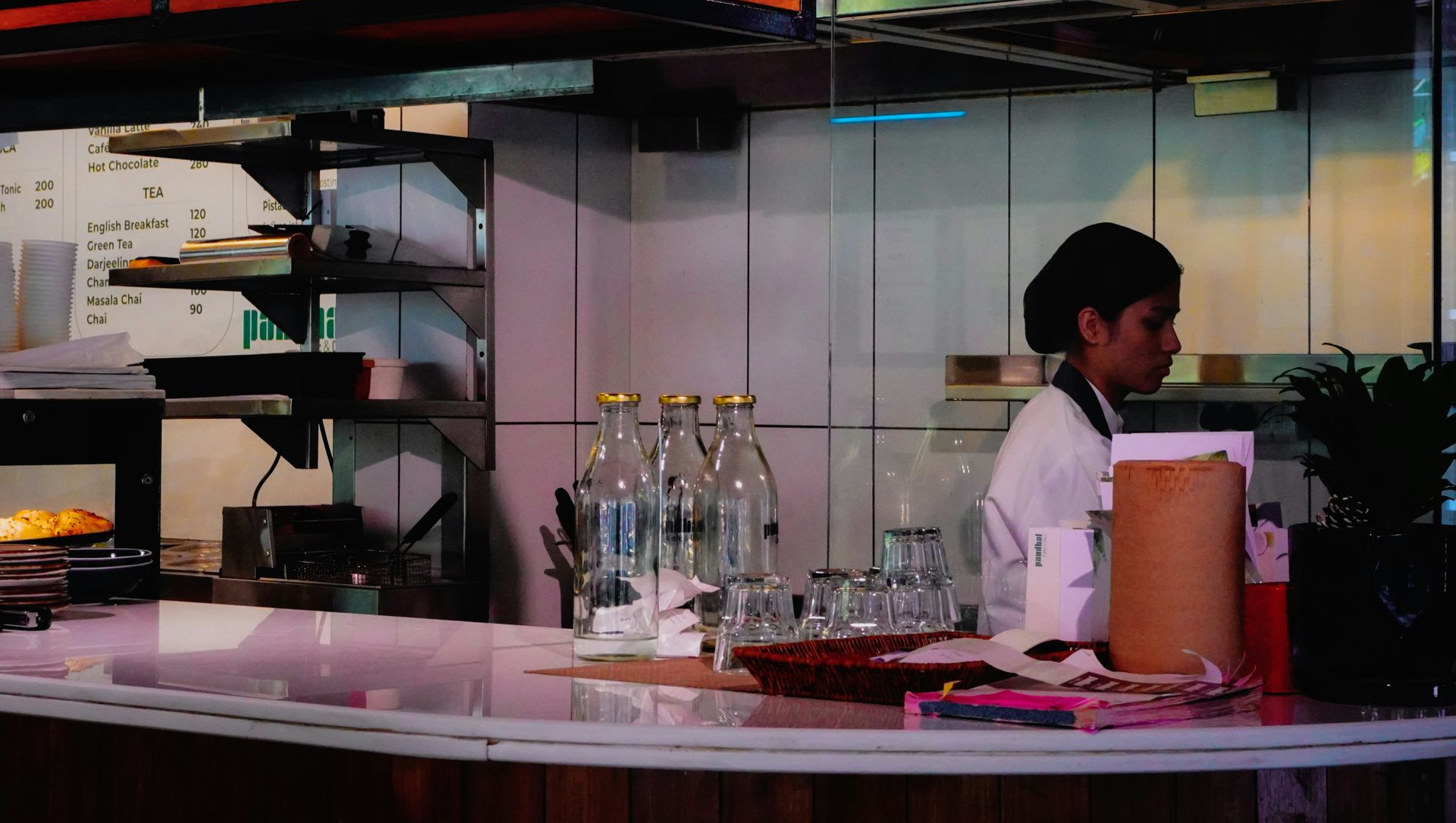 A restaurant employee stands behind a service counter with glass bottles and drinking glasses in the foreground.