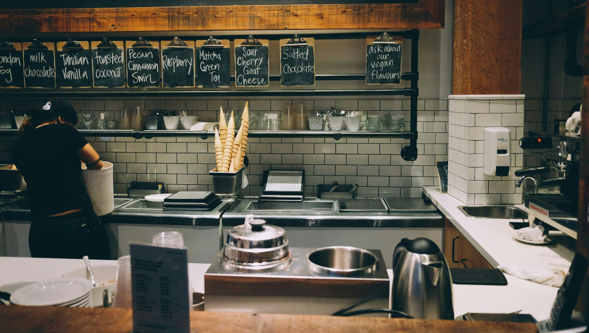 Cozy café counter with chalkboard menu, shelves, pastries, coffee machine, and a barista working behind the bar