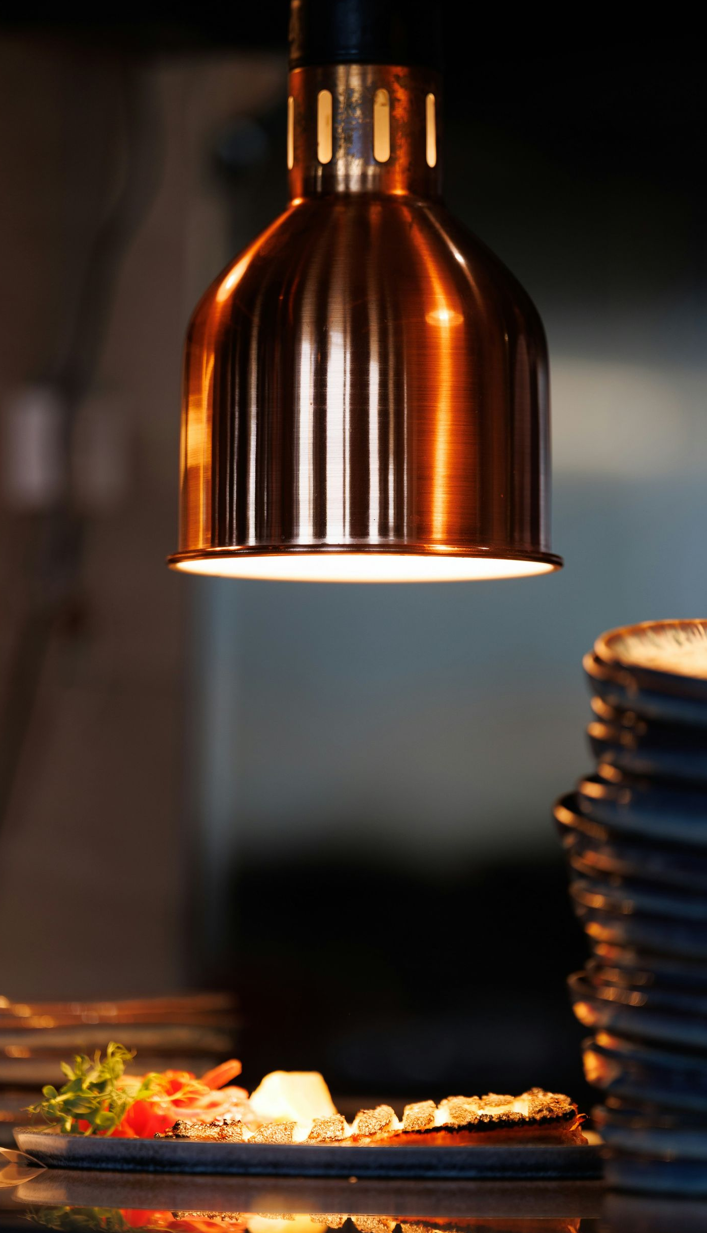 A copper heat lamp hangs above a plated dish and a stack of ceramic bowls in a dimly lit restaurant kitchen.