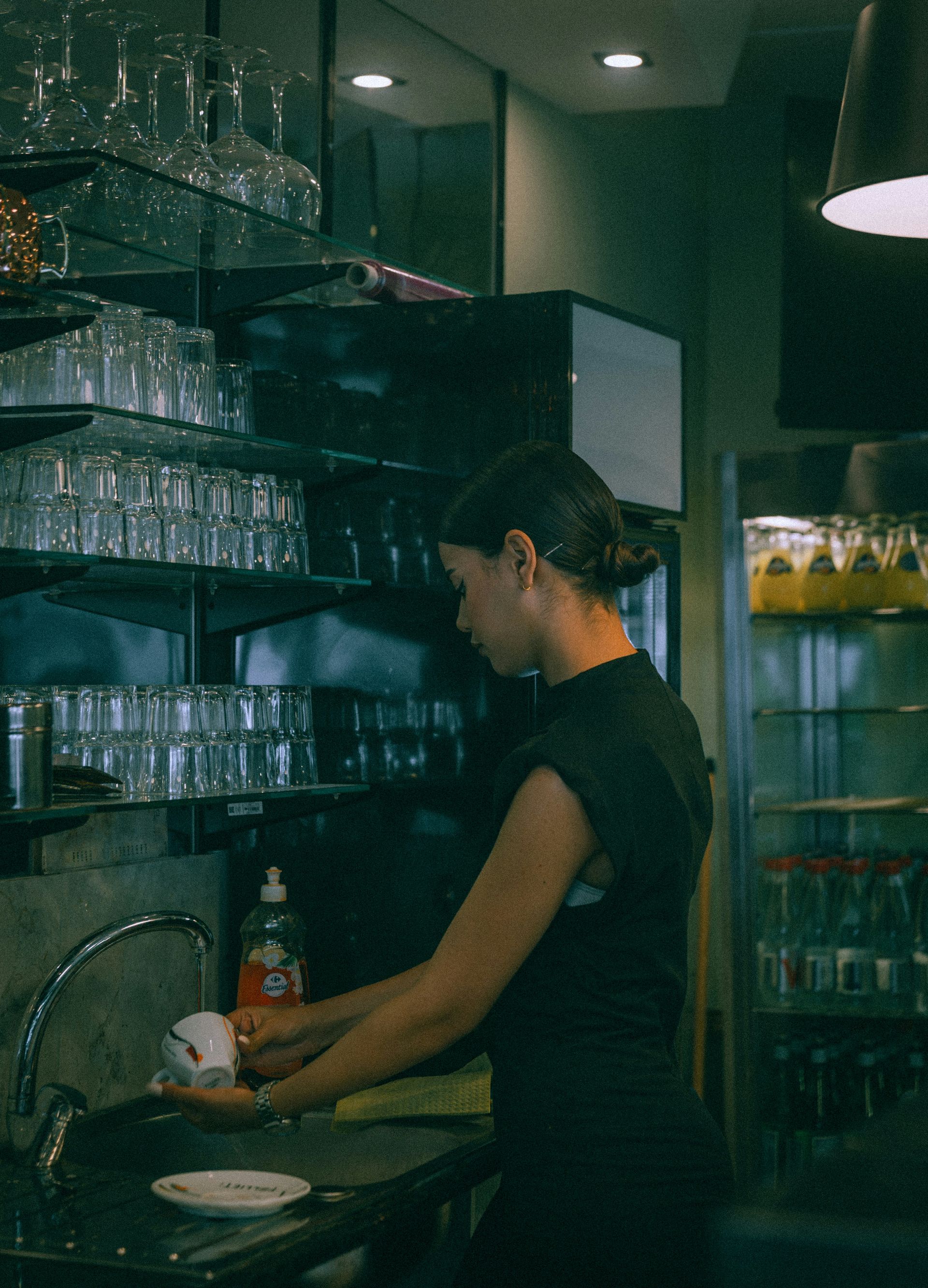 Person washing dishes at a restaurant sink beside shelves of glassware and bottles.