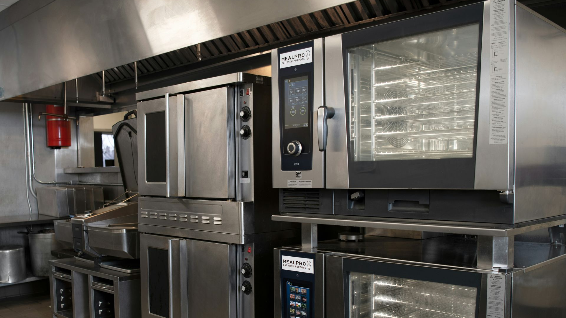 Stainless steel commercial ovens stacked in a professional kitchen under a metal hood.