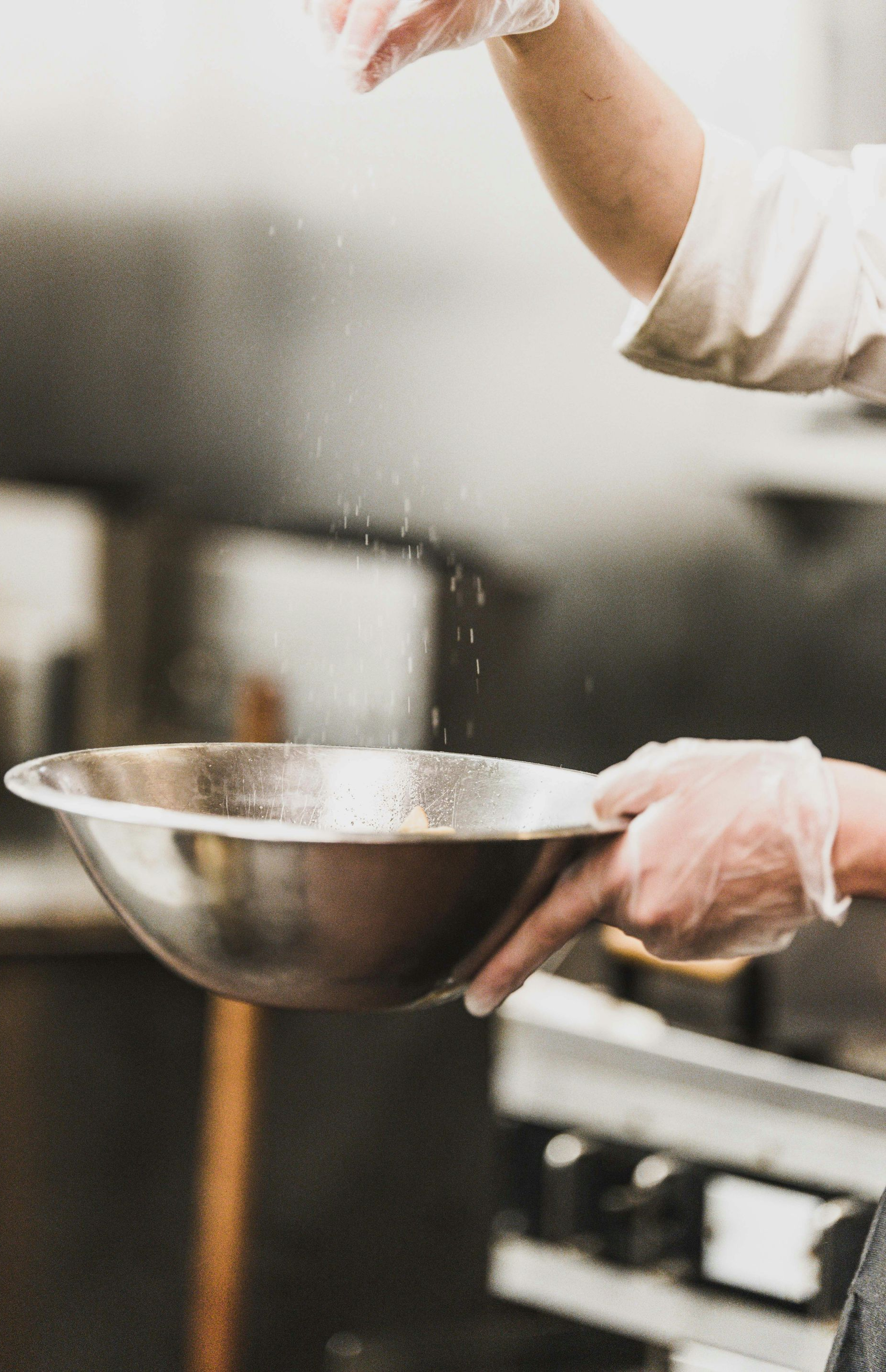 Person holding a metal mixing bowl in a kitchen, sprinkling ingredients into it.