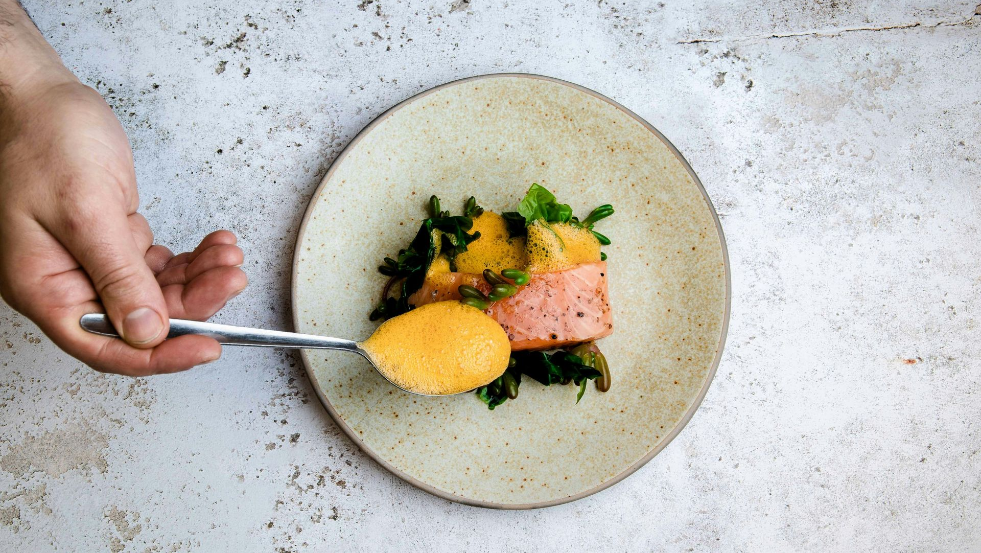 Hand spooning orange sauce onto a plated dish with greens and pink fish on a stone table