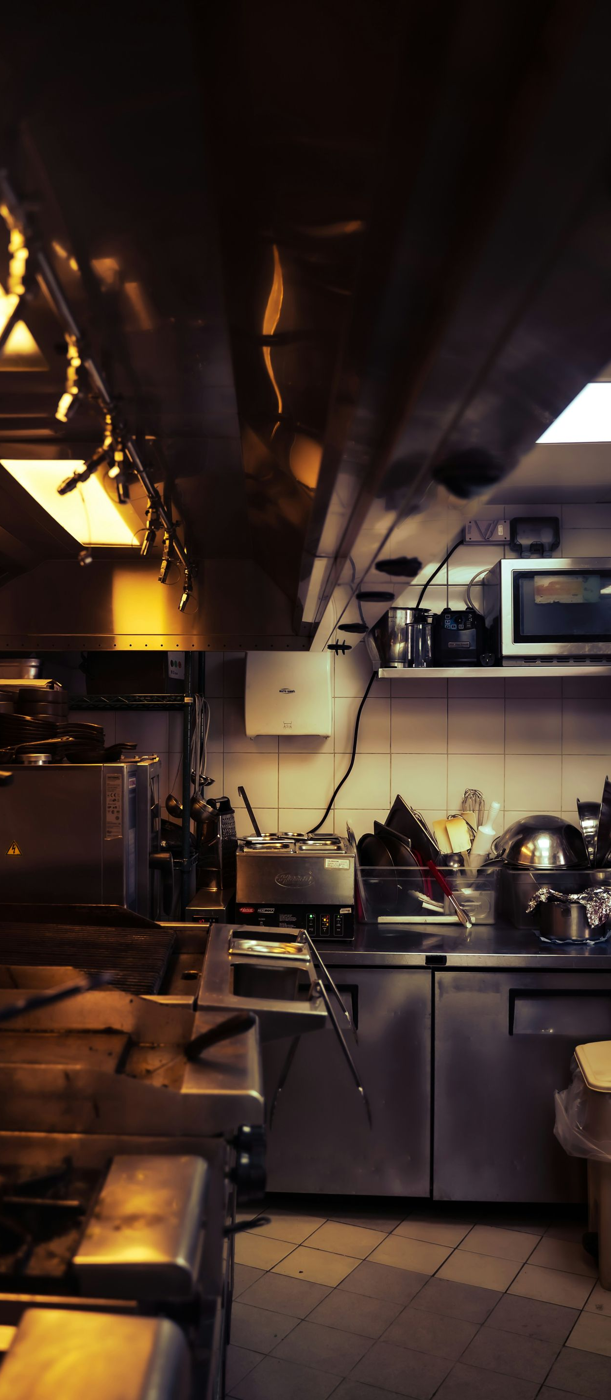 Dimly lit industrial kitchen with stainless steel counters, pots, and hanging utensils under warm lights