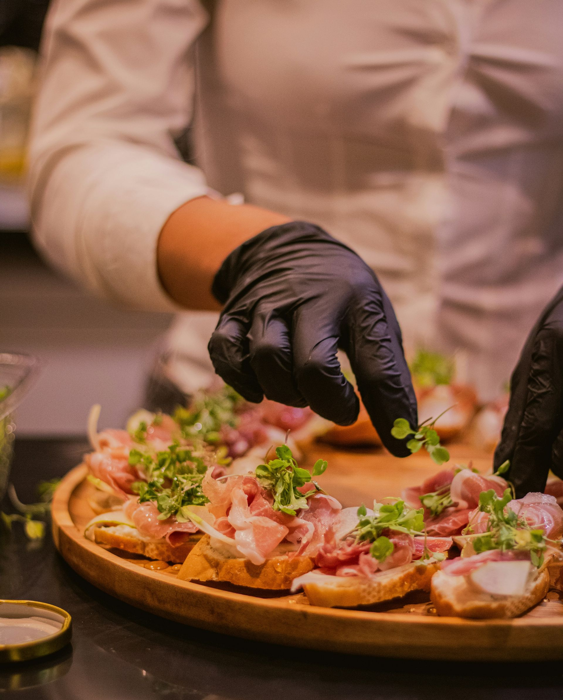 Chef in black gloves garnishing a wood-fired pizza on a wooden board with fresh basil and meat slices