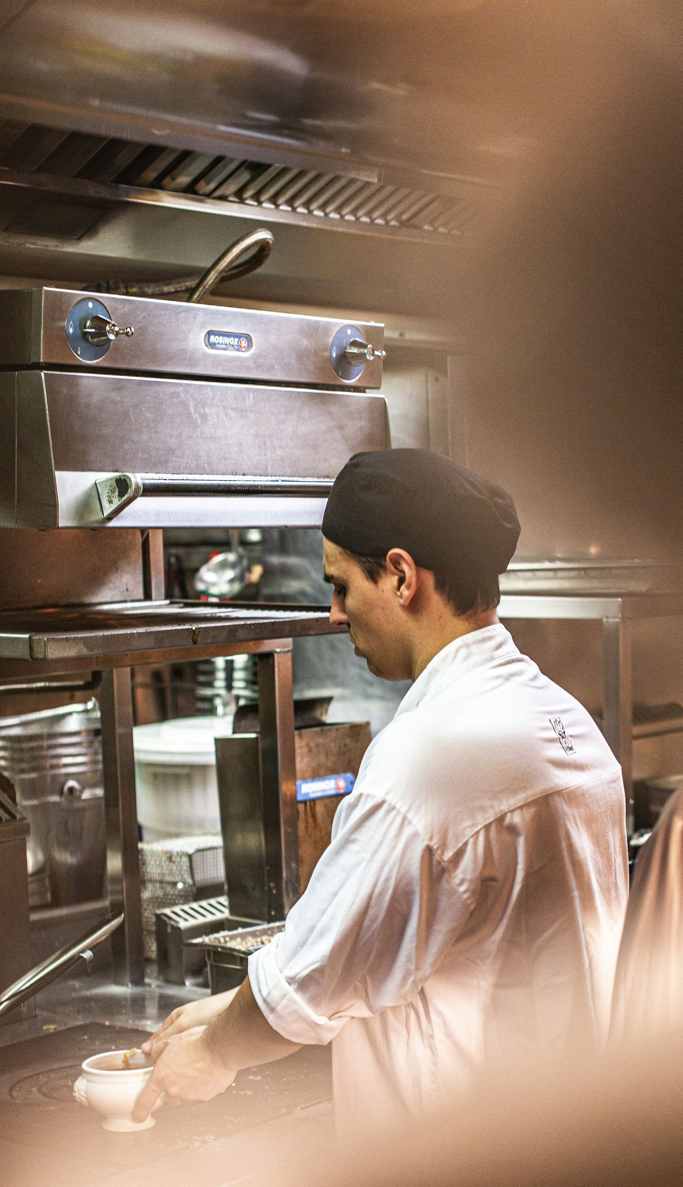 Chef in white jacket working at a stainless-steel kitchen counter, seen from behind.