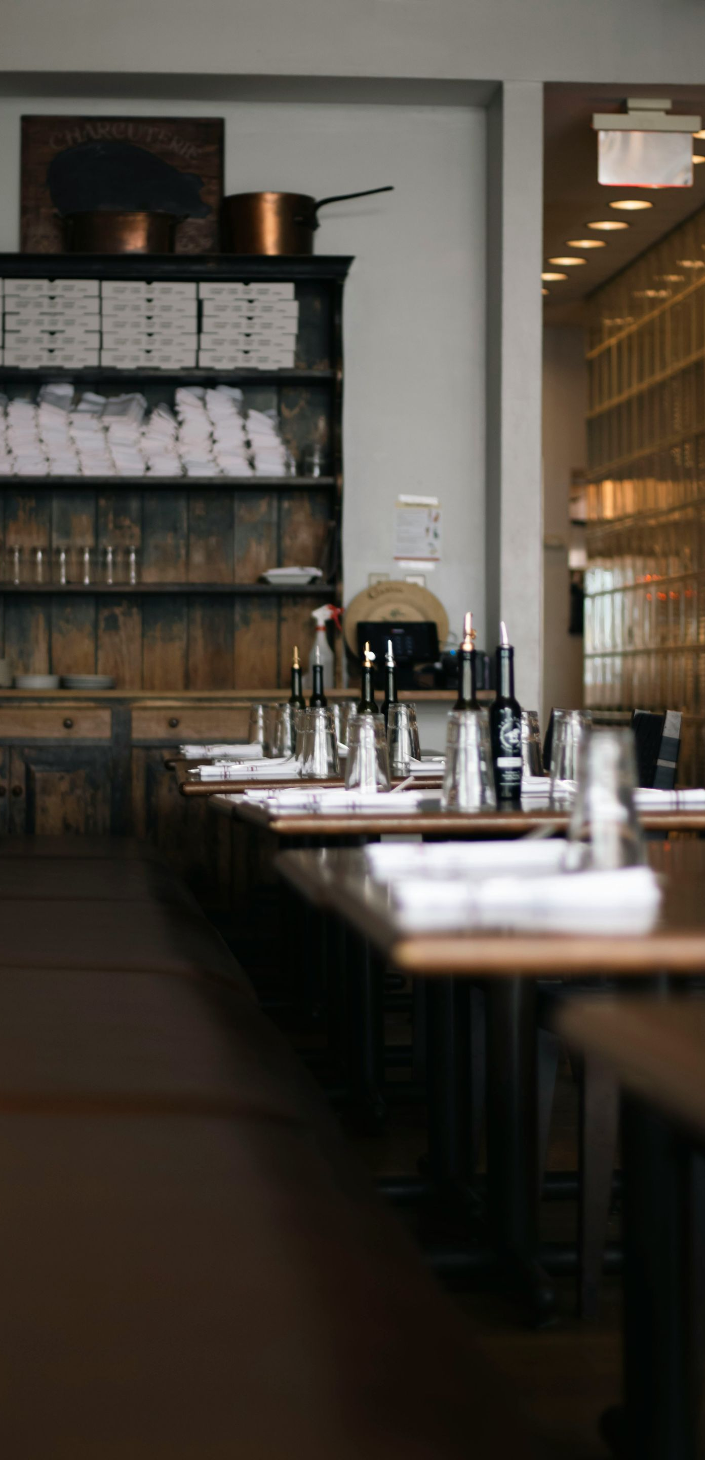 An empty restaurant dining area with set tables, wooden benches, and a dark shelving unit filled with white linens.