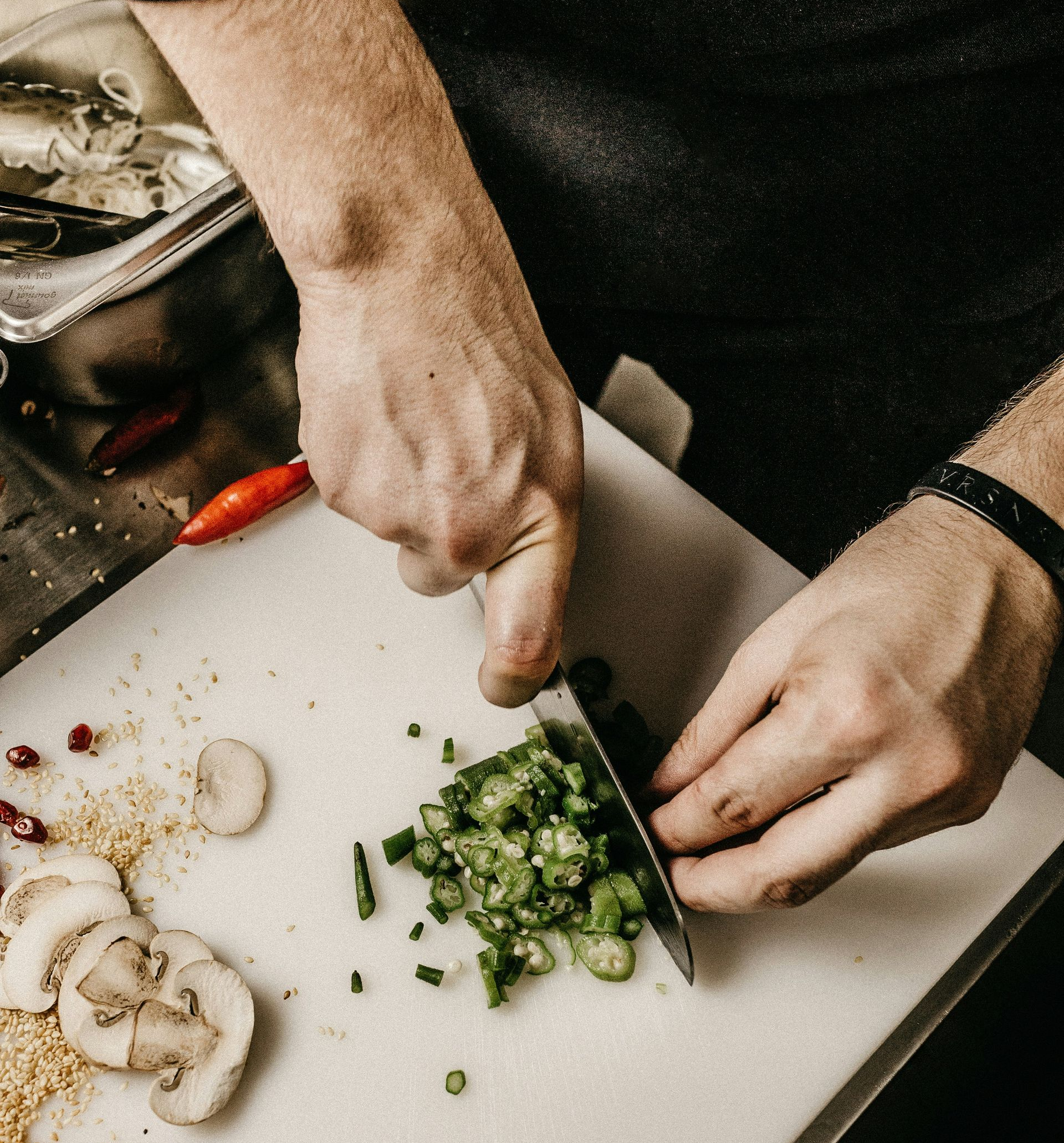 A person’s hands use a knife to finely chop green jalapeño peppers on a white cutting board near sliced mushrooms.