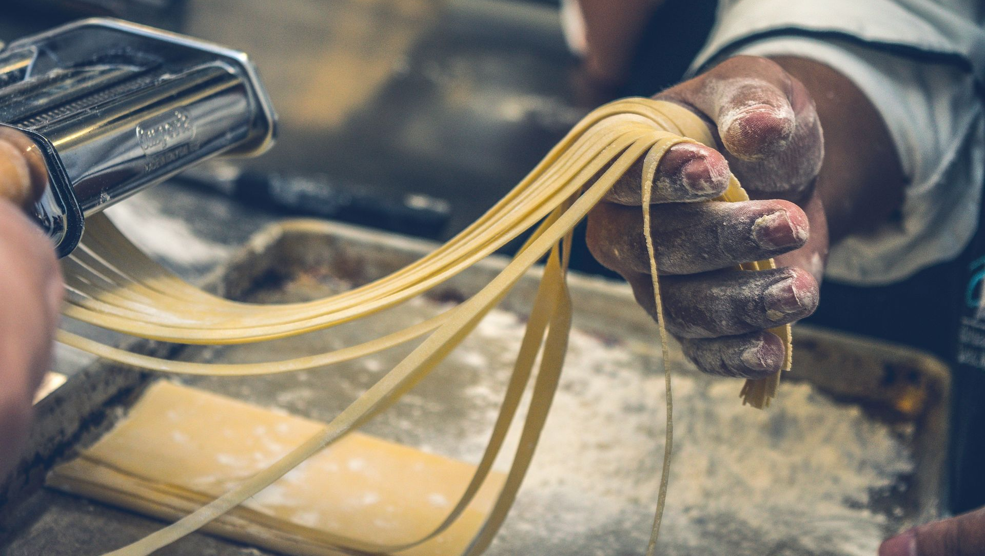 Chef making pasta with a pasta machine, holding freshly cut noodles dusted with flour.