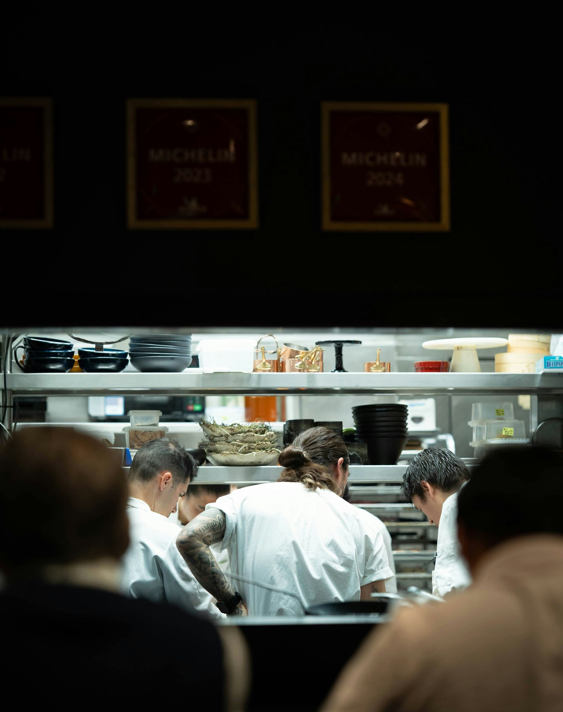 People standing at a brightly lit restaurant kitchen counter, viewed through a glass window.
