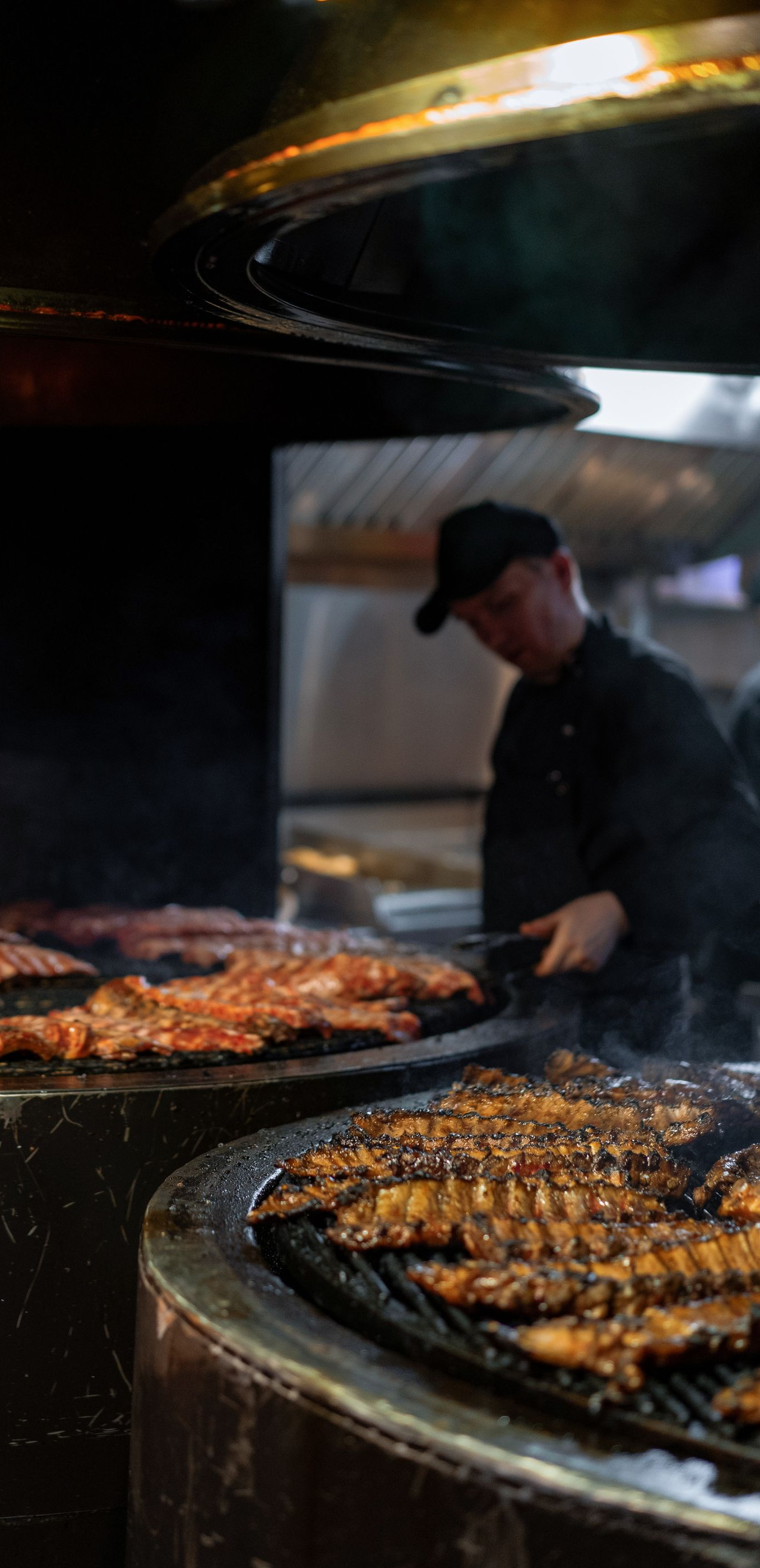 Chef grilling meat on large circular barbecue pits in a smoky kitchen