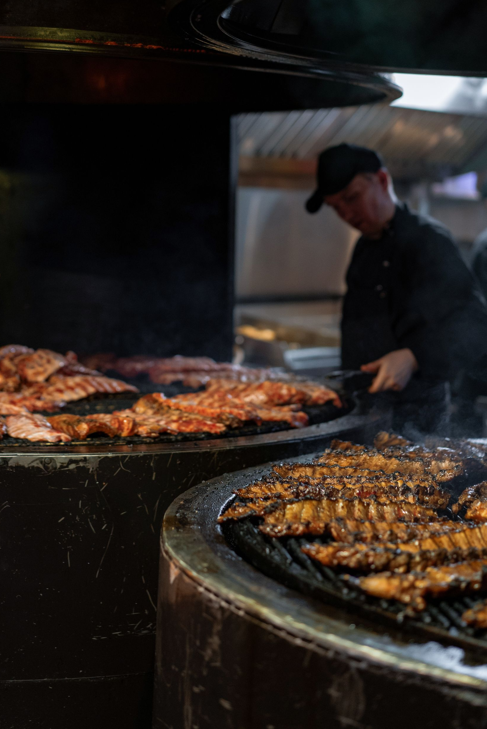 Chef grilling meat on a large charcoal barbecue, with skewers and steaks sizzling over open heat.