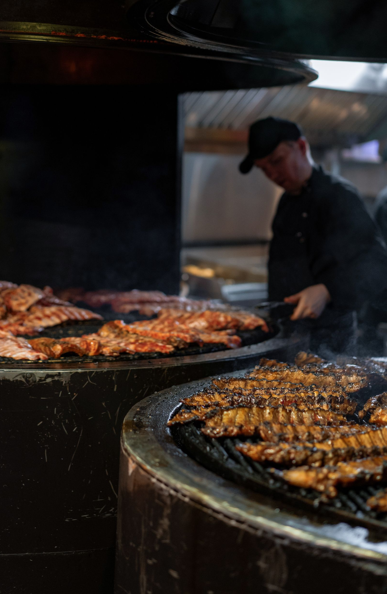 Chef grilling meat on a large charcoal barbecue, with skewers and steaks sizzling over open heat.