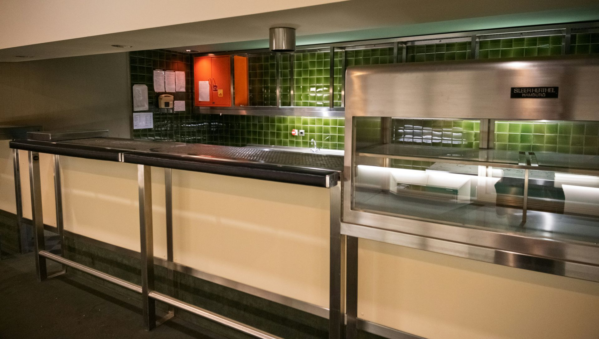 A stainless steel food service area with a warming oven and a long counter. Green tiled wall backdrop.