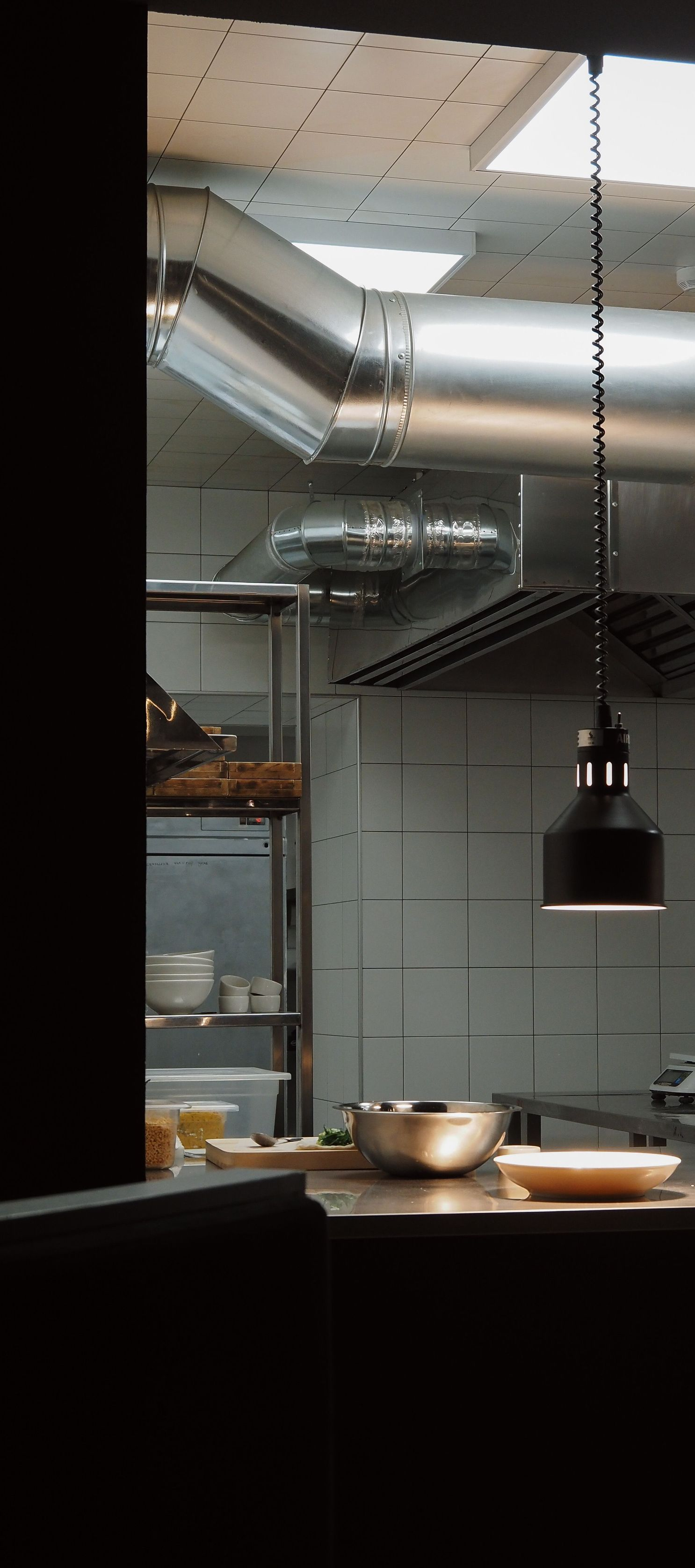 Commercial kitchen with stainless steel vents, hanging heat lamp, and pots on a counter under bright ceiling lights