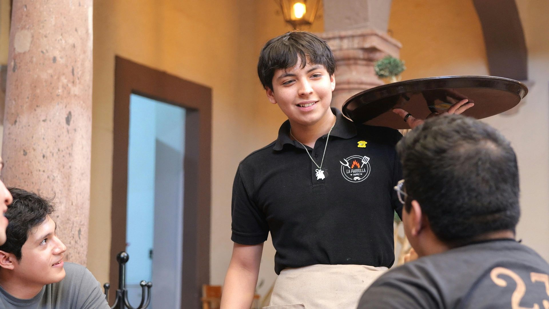 Server holding tray, smiling at patrons in an outdoor restaurant setting.