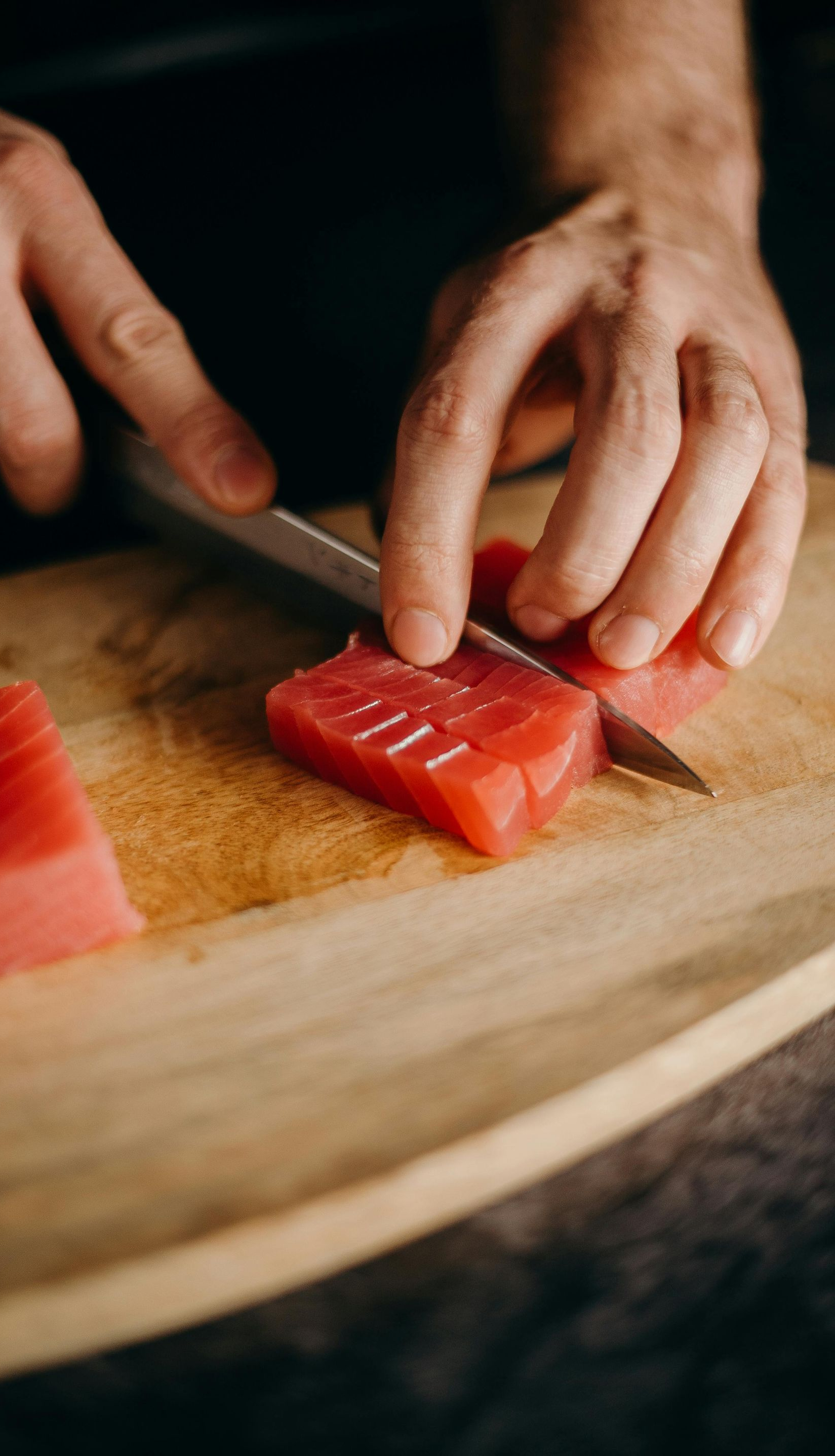 Hands slicing red tuna on a wooden cutting board with a silver knife.