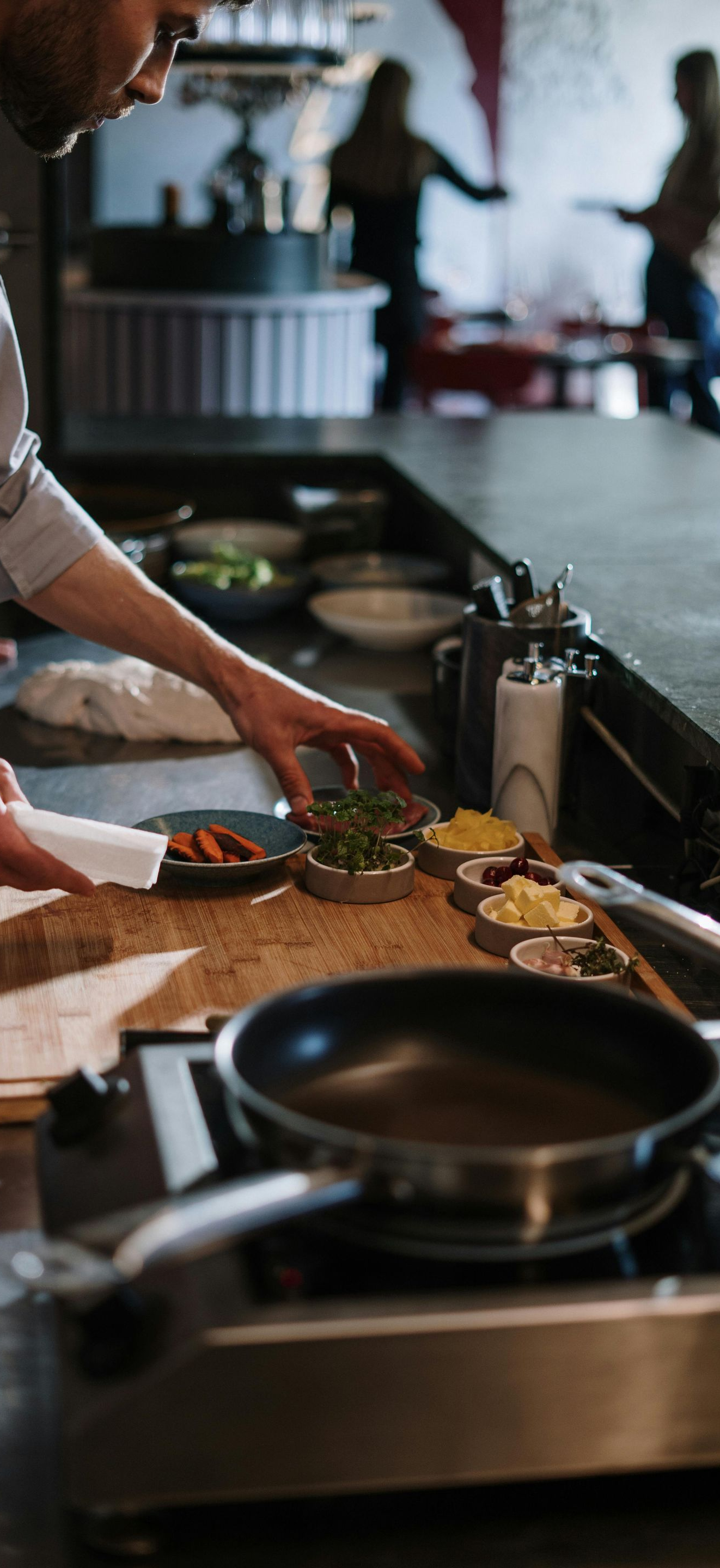A person adds fresh herbs to small garnish bowls on a kitchen counter in a restaurant, with an empty pan on the stove.