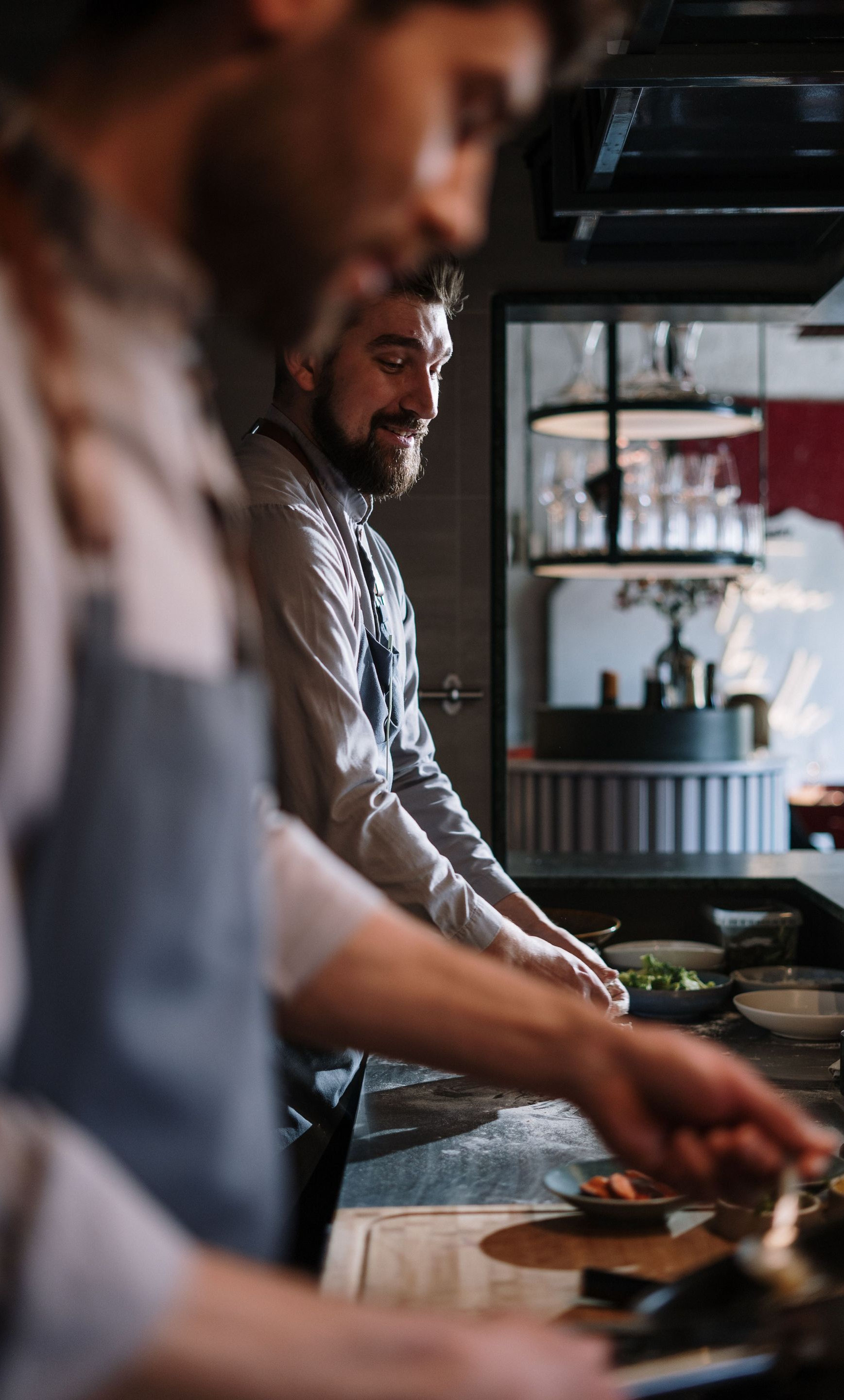 Chefs preparing food in a busy restaurant kitchen with stainless steel counters and hanging shelves