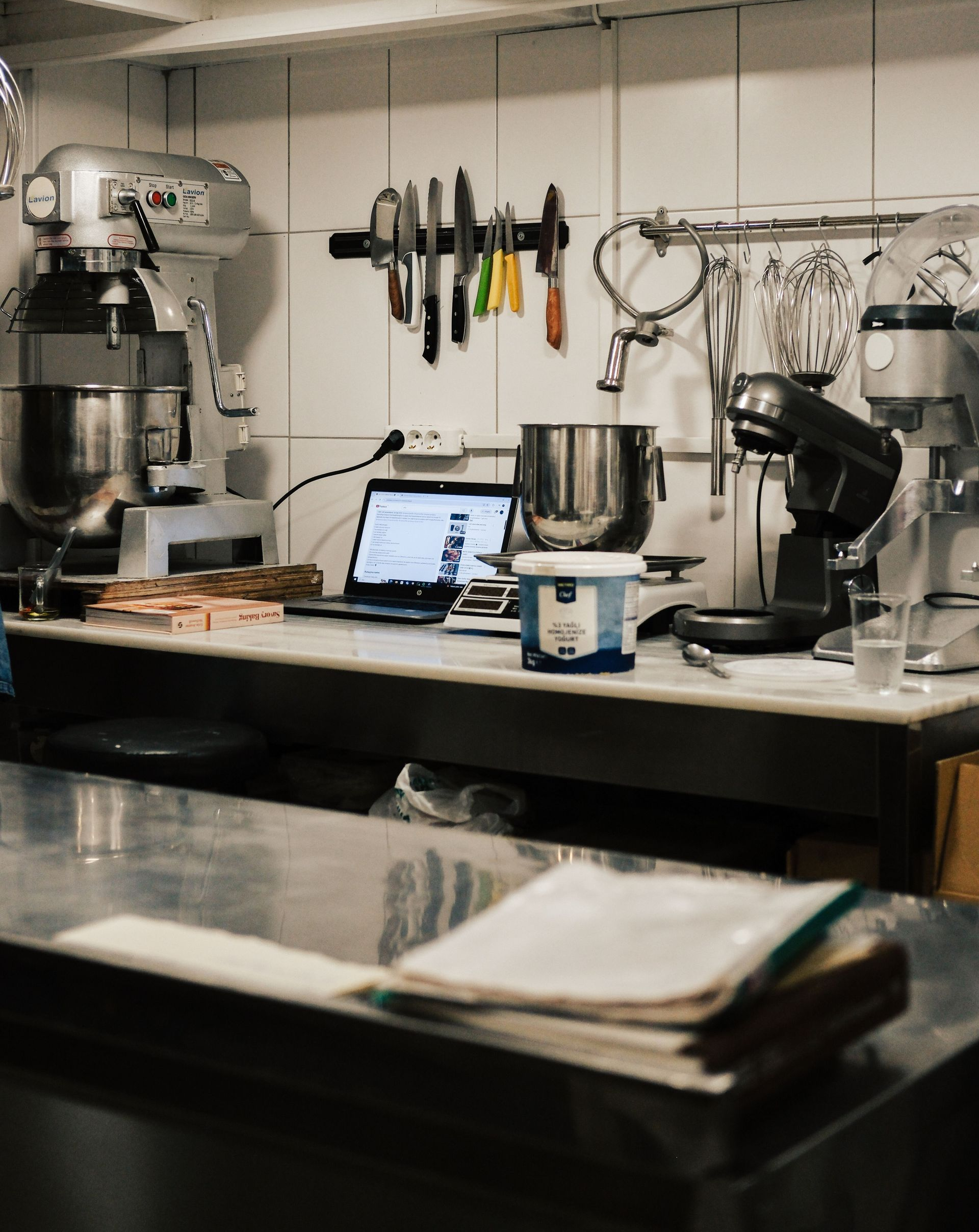 Coffee shop prep counter with espresso machine, tools, laptop, and mugs under bright overhead lights