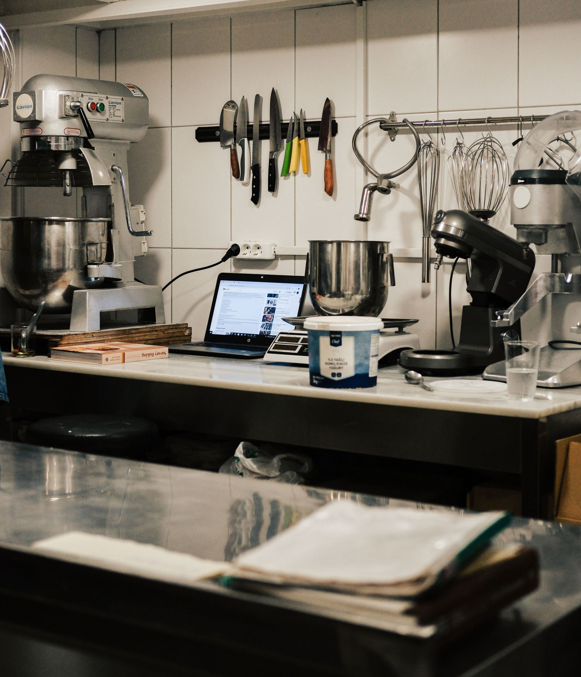 Commercial kitchen counter with mixers, utensils, a laptop, and metal bowls on a work surface.
