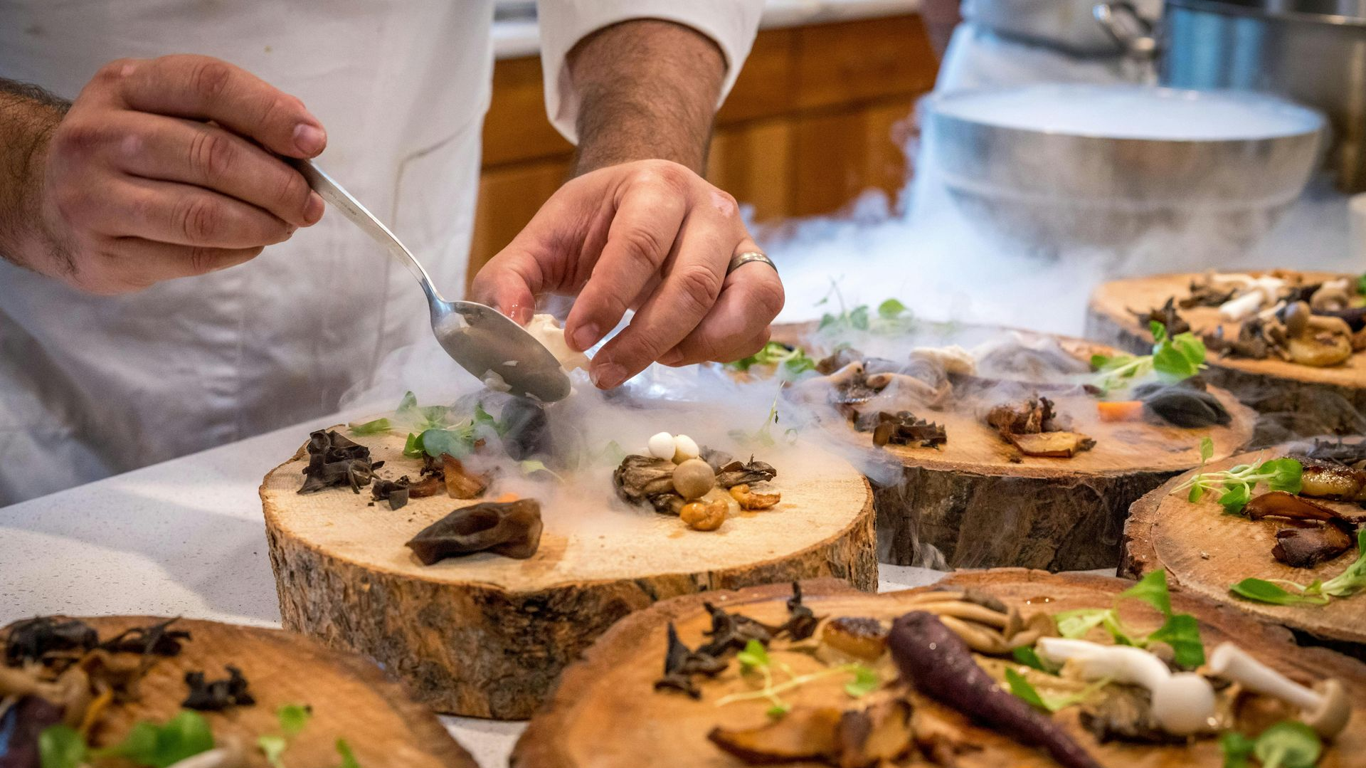 Chef plates mushroom appetizer on wooden rounds, using a spoon with dry ice.