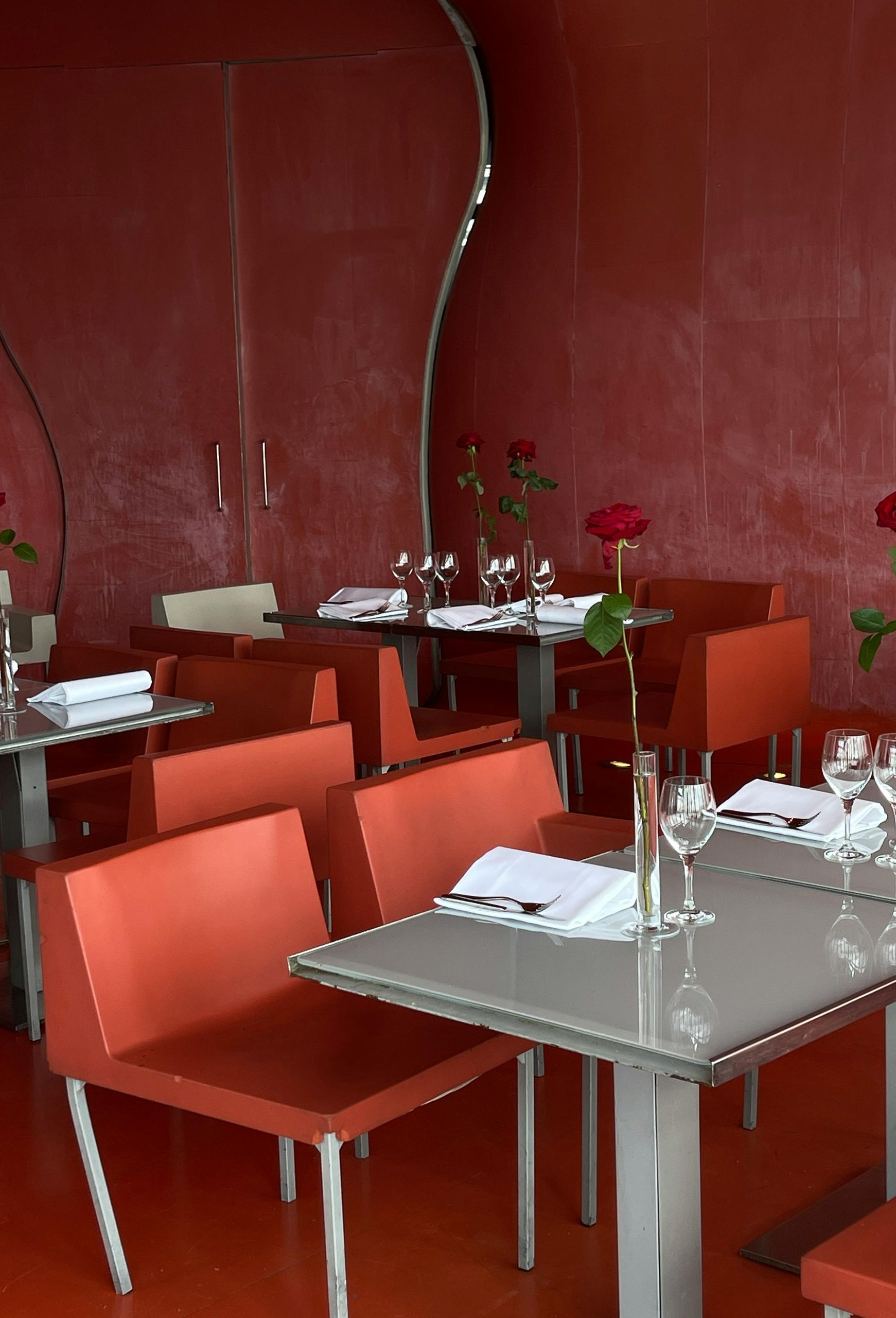 Red-toned restaurant interior with orange seating, gray tables set with glasses and white napkins, and single red roses in vases.