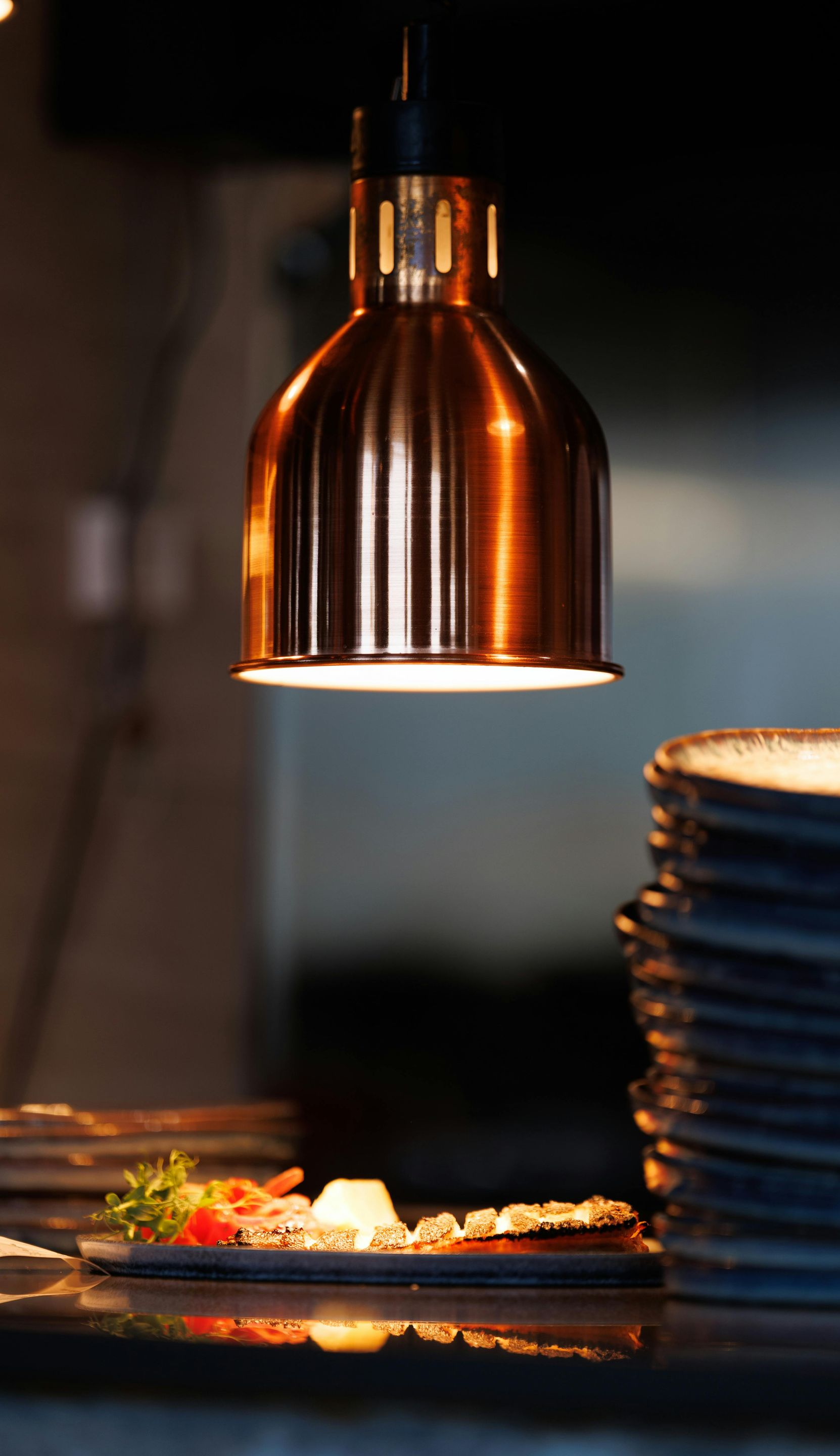 Warm-lit copper pendant lamp over a plated gourmet dish beside stacked plates in a dim restaurant setting