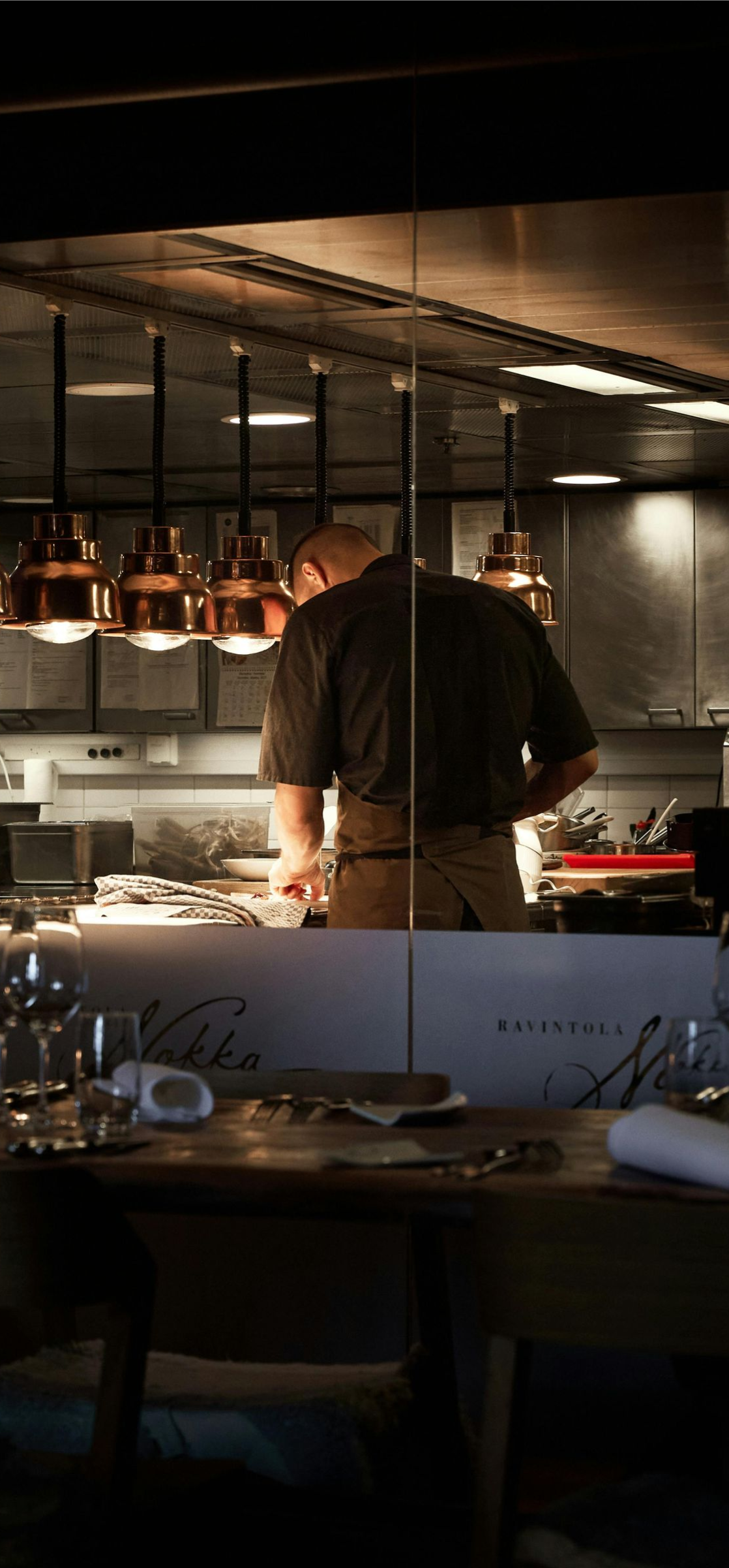 Chef working under warm lights in a restaurant kitchen behind a dining table set with glasses and plates