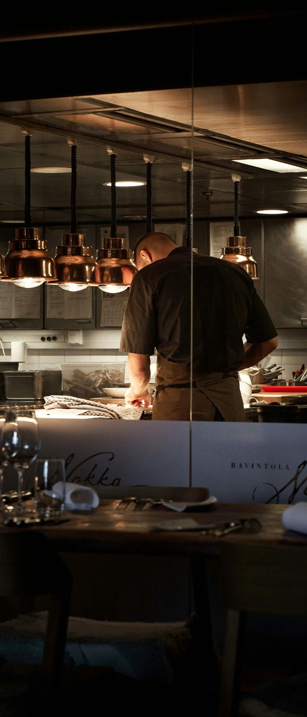 Chef working at a dimly lit restaurant kitchen counter with hanging copper pans above.