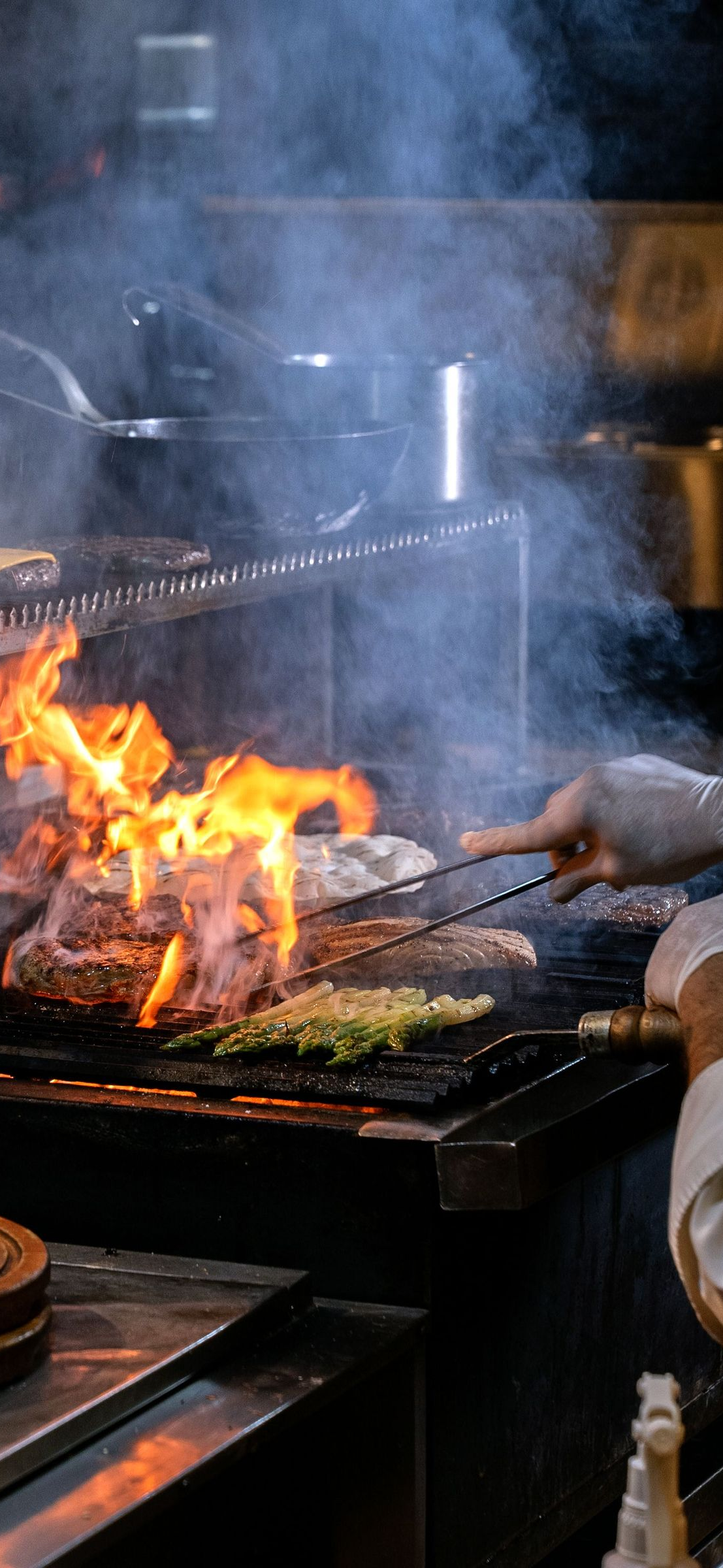 Steak and vegetables sizzling on a smoky outdoor grill with flames rising.