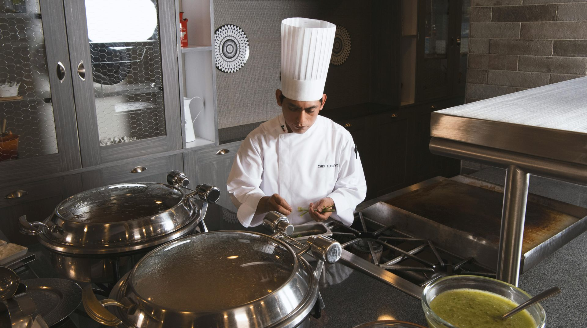 A chef in uniform prepares food behind a counter with silver serving dishes and bowls of sauce in a kitchen setting.