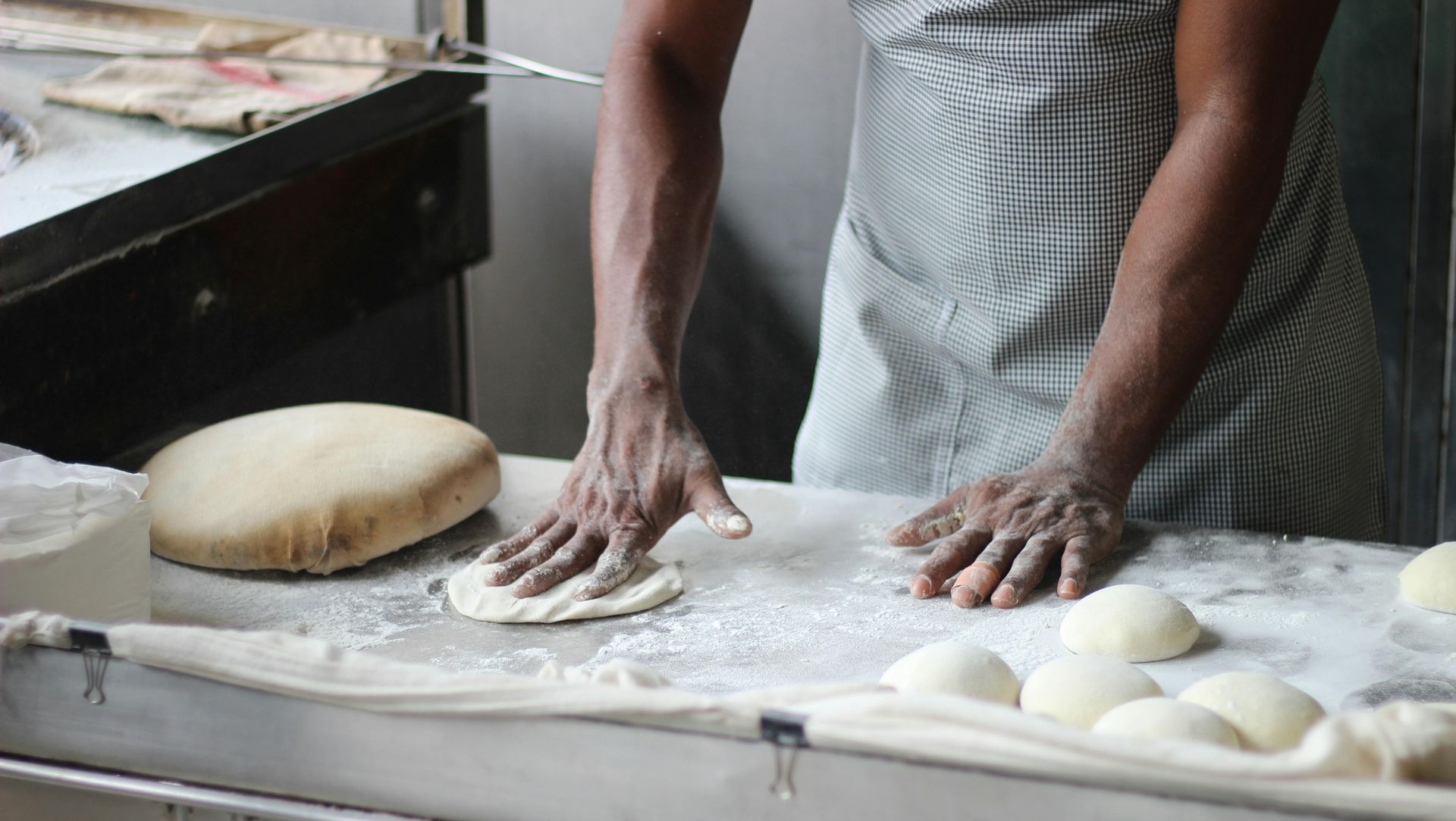 Person flattening dough on a floured surface, with loaves and a conveyor belt.