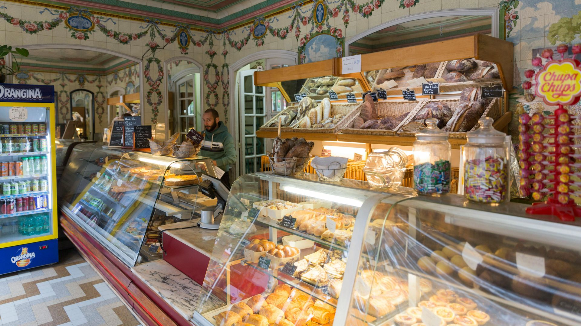 Bakery interior: display cases filled with pastries, ornate floral wallpaper, person behind the counter.