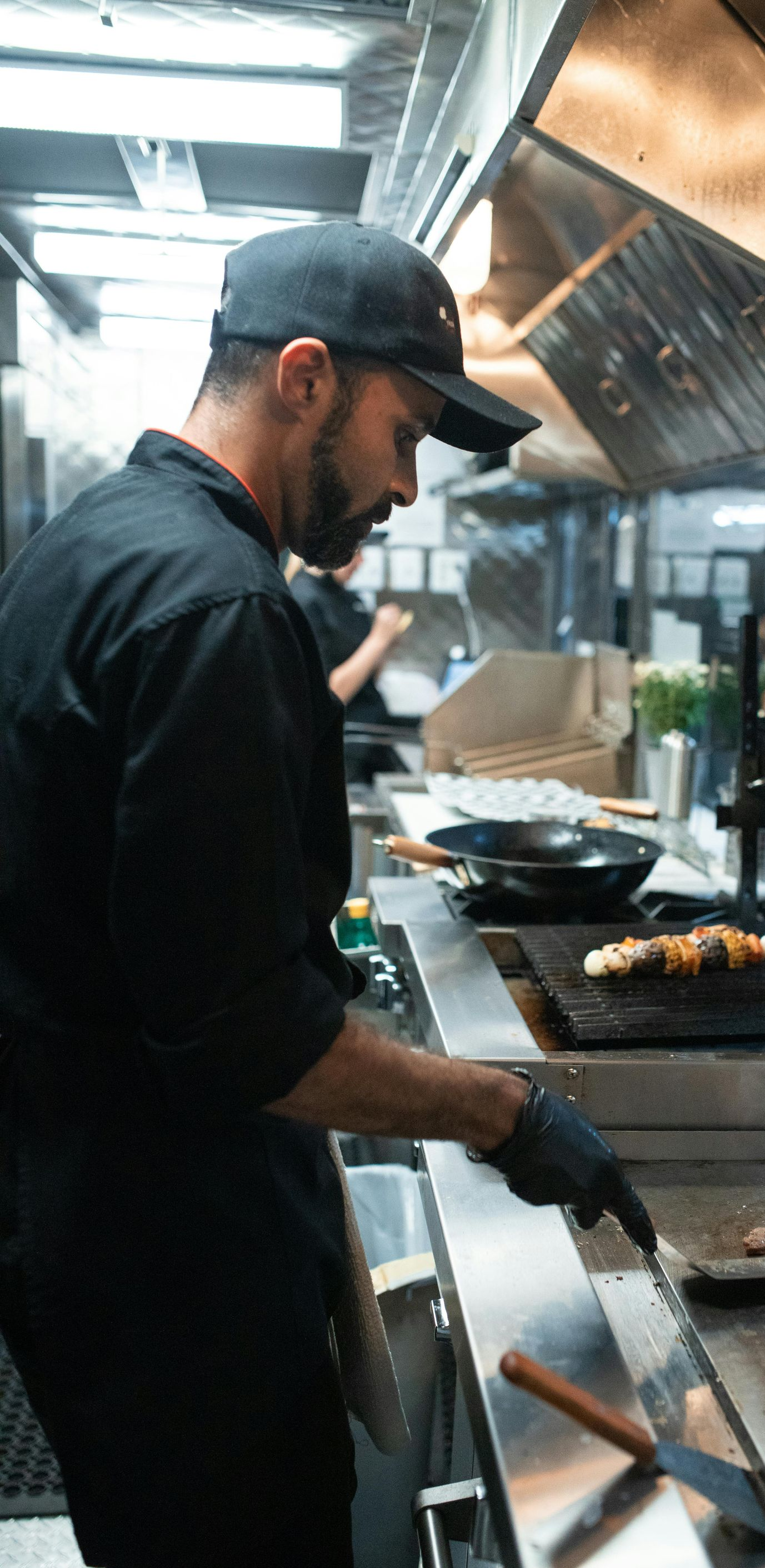 A chef in a black uniform and cap flips food on a griddle in a commercial kitchen.